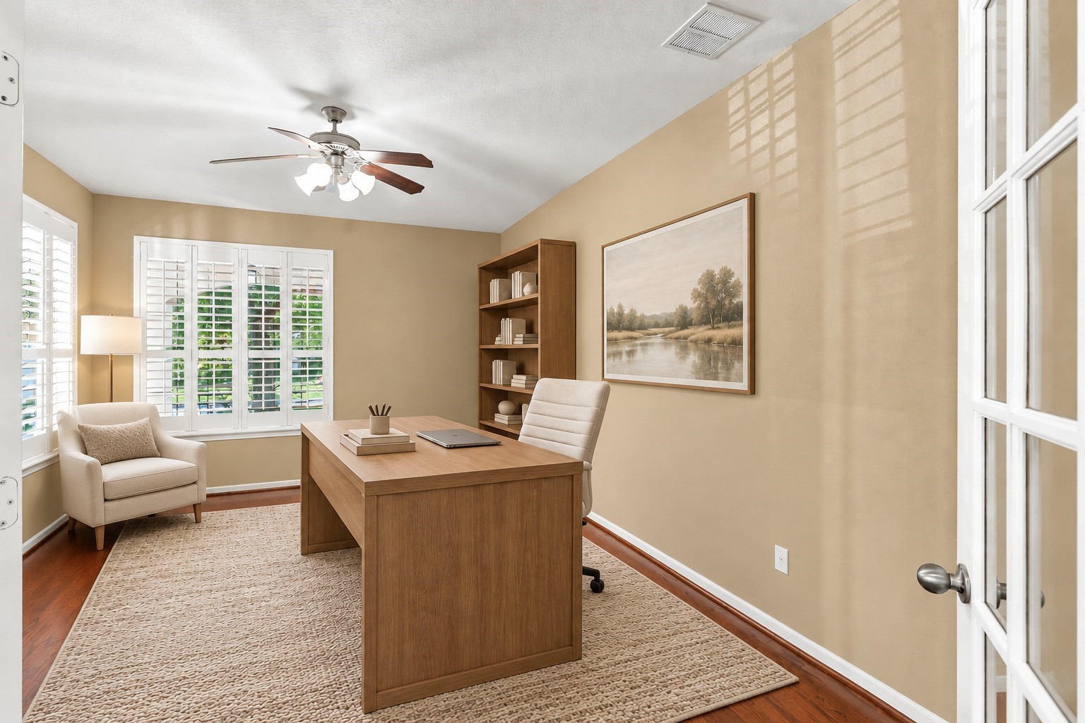 Virtually Staged: A bright and inviting home office featuring warm neutral walls, rich hardwood flooring, and abundant natural light from multiple windows with plantation shutters. A ceiling fan with integrated lighting provides added comfort, and the spacious layout offers plenty of room for a functional and comfortable workspace.