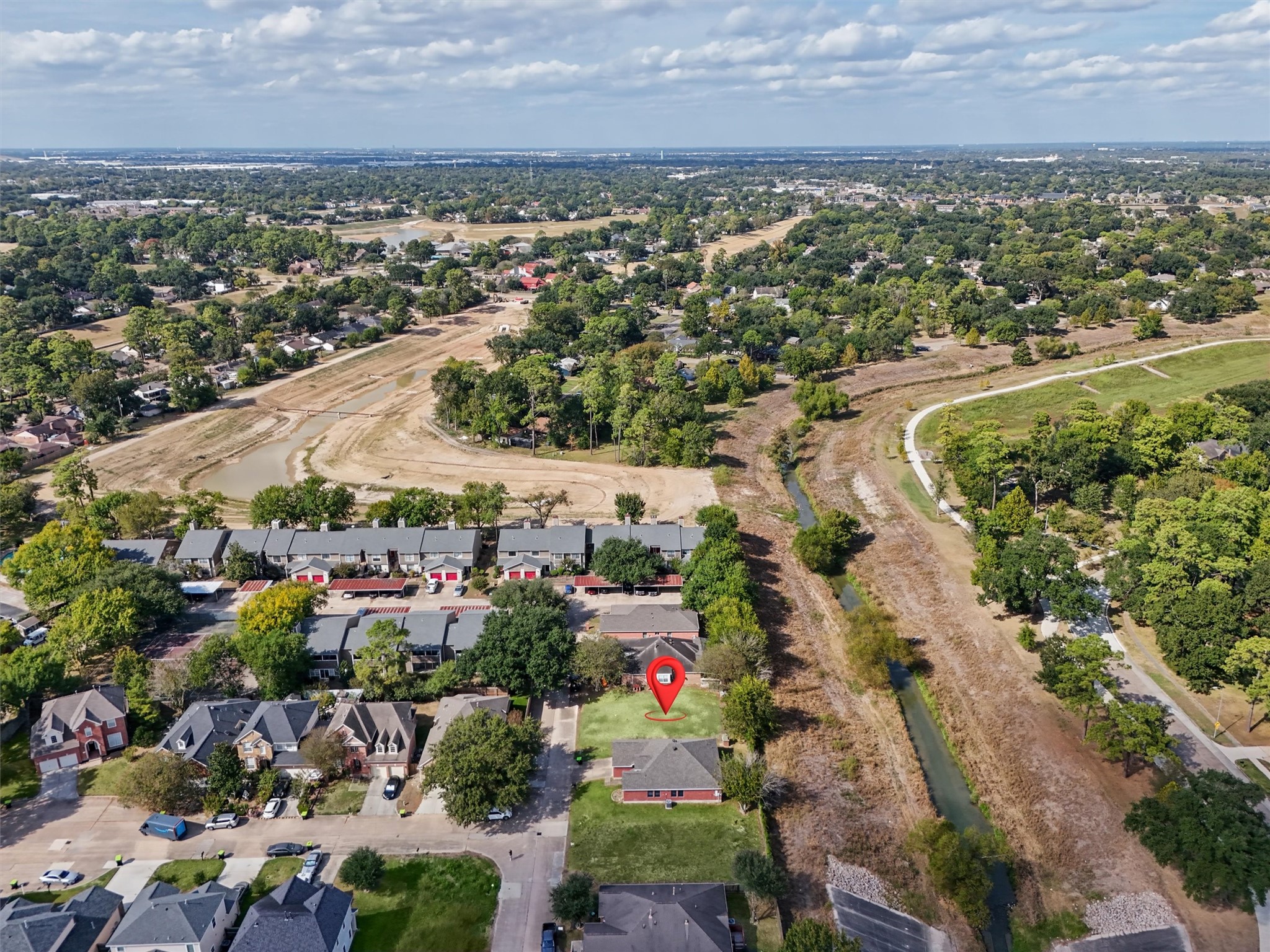 The red arrow highlights this cleared residential lot, surrounded by established homes, mature trees, and beautiful green spaces. This aerial view showcases the nearby walking trails, winding waterway, and expansive open areas that add to the neighborhood's charm. Enjoy the convenience of nearby shopping, dining, and everyday amenities while creating your own custom dream home in this inviting residential setting.