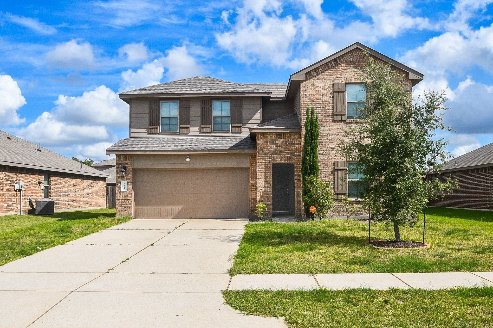 Charming two-story brick home with a spacious driveway and attached garage, set against a lush green lawn and clear blue skies.