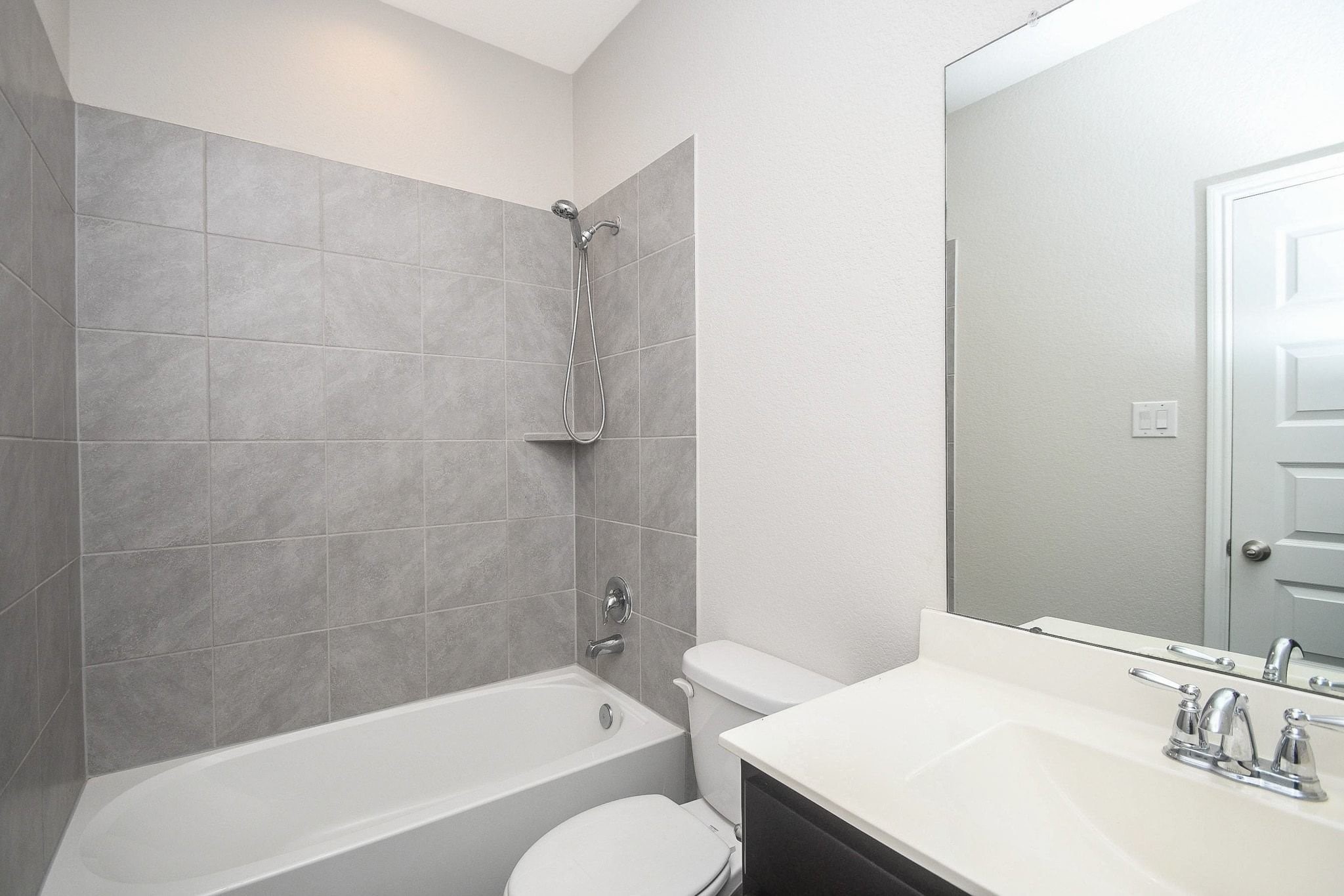 Modern bathroom downstairs featuring sleek gray tile, a pristine bathtub with shower, and a spacious vanity with a large mirror for a clean and contemporary look next to the secondary bedroom.