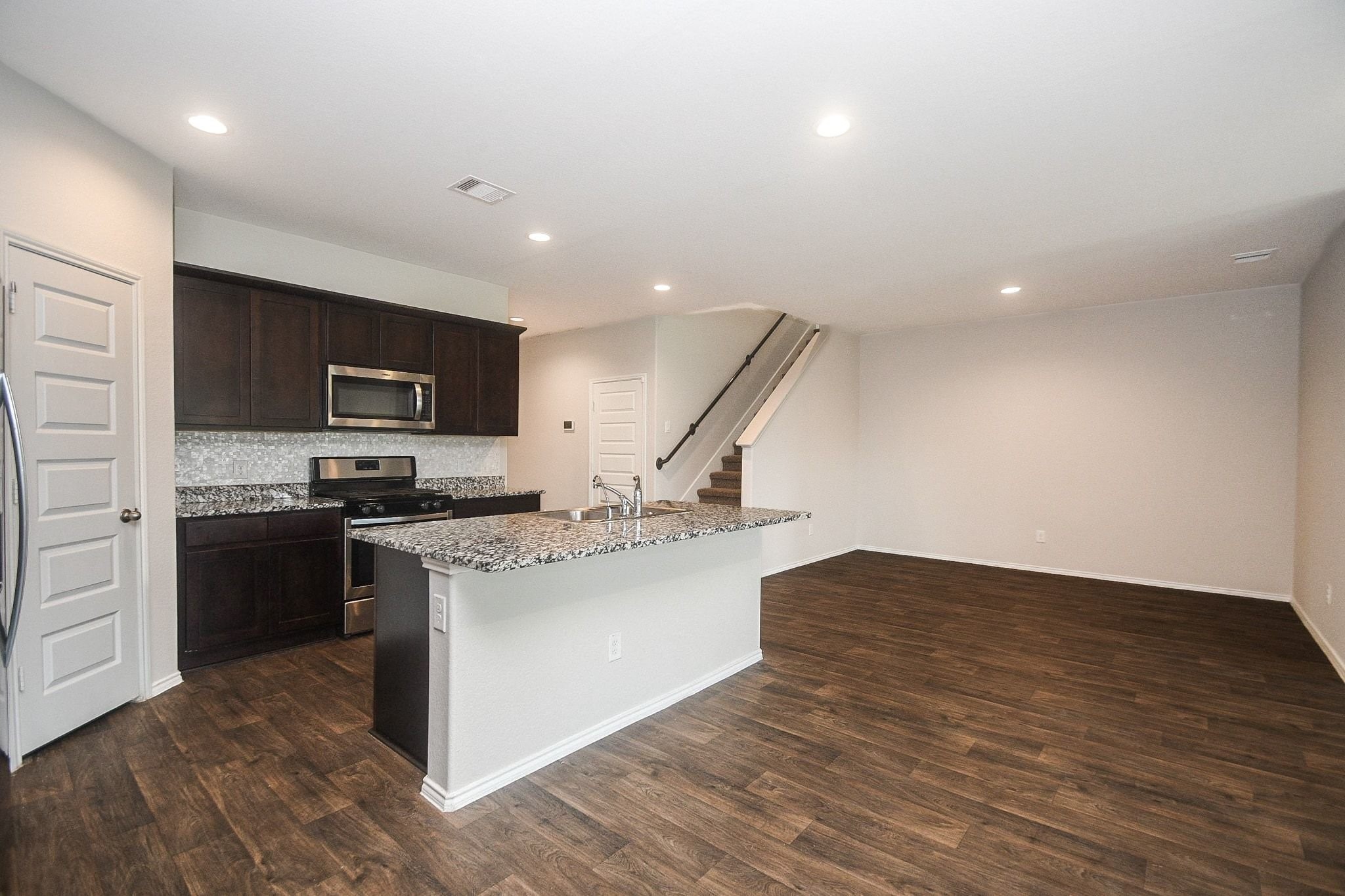Open-concept living area with a modern kitchen featuring dark cabinetry, stainless steel appliances, and a granite island, complemented by warm wood flooring and recessed lighting with a view of the dining or study area.