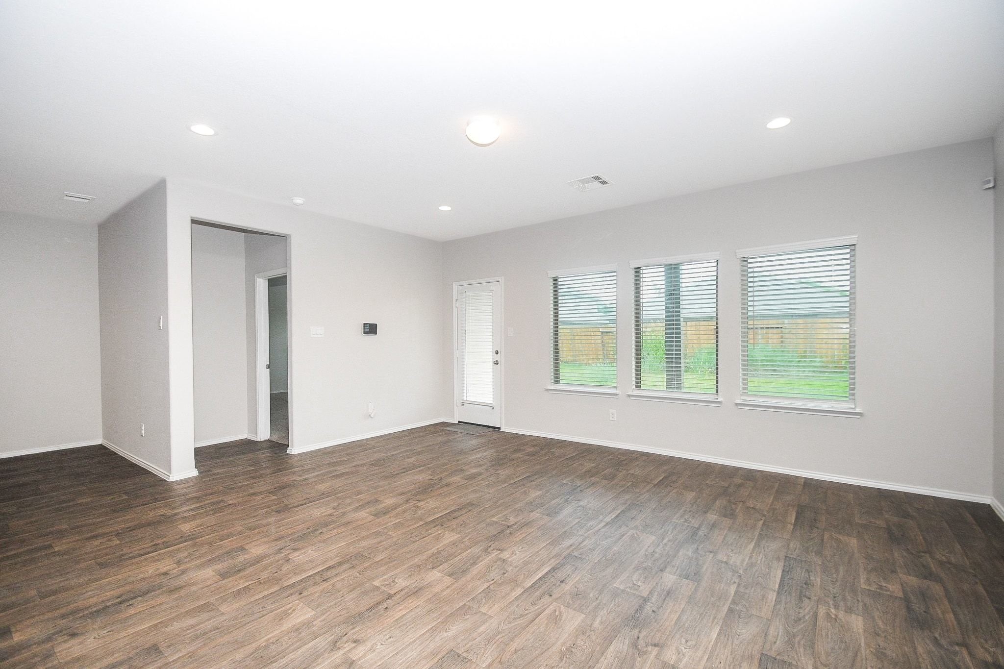 Bright and spacious living area with rich hardwood flooring, large windows offering plenty of natural light, and a door leading to the backyard and showing entry to the primary suite downstairs.