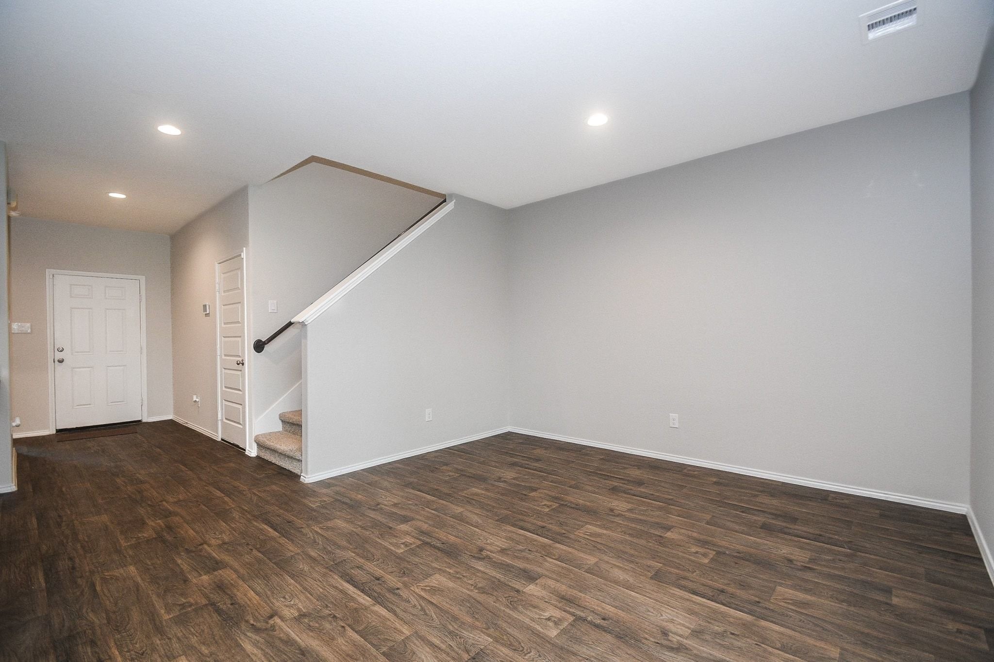 Spacious living area with elegant wood flooring, neutral walls, and recessed lighting, featuring an inviting staircase and convenient entryway.