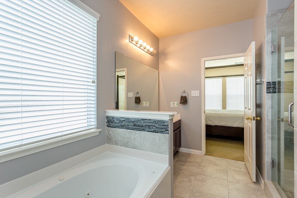 Experience relaxation in this elegant bathroom featuring a spacious soaking tub, modern vanity with stylish tile accents, and a glass-enclosed shower. Natural light filters through the large window, enhancing the serene ambiance.