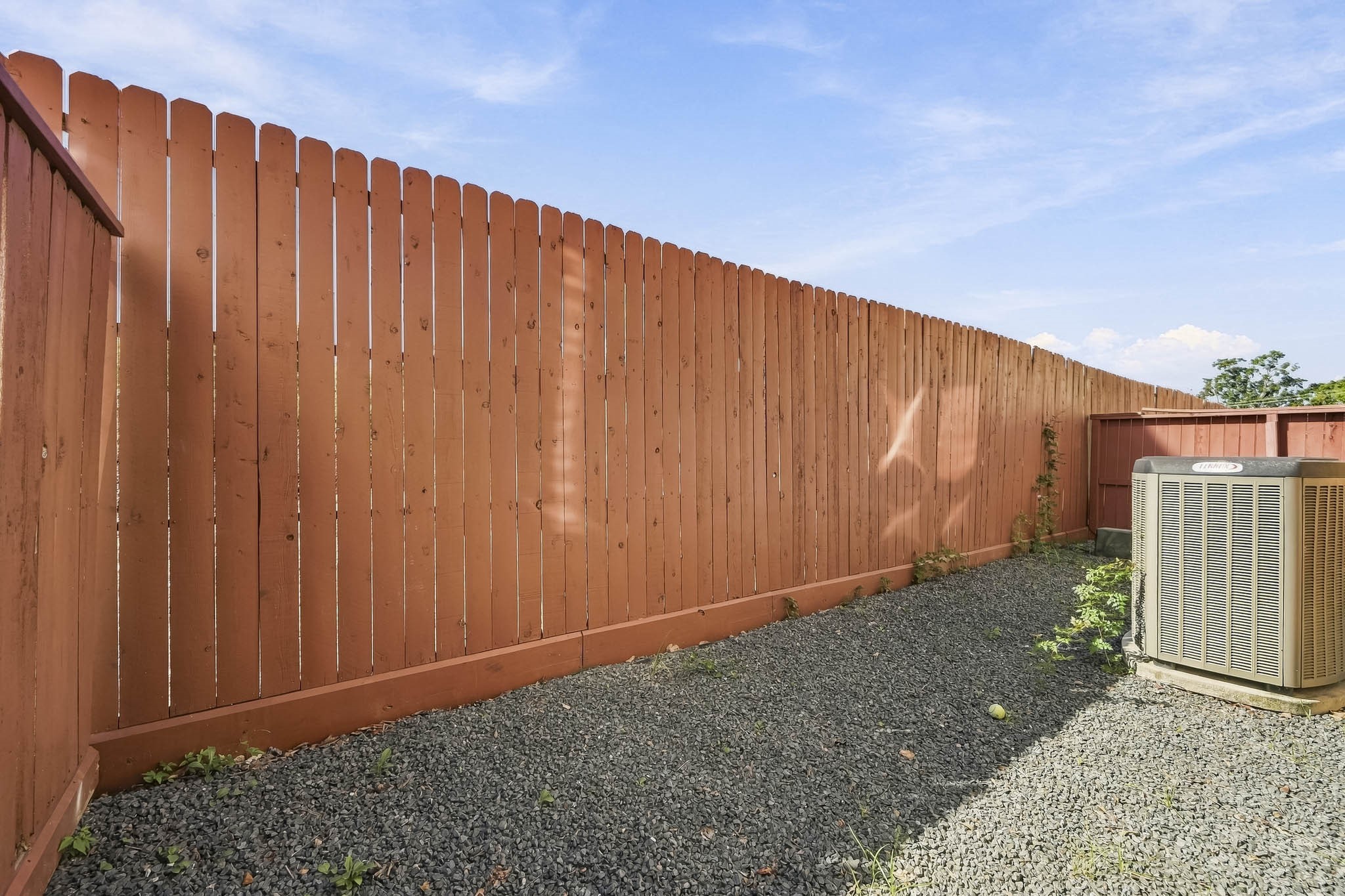 Private outdoor space with a tall wooden fence, gravel ground, and an efficient air conditioning unit, perfect for low-maintenance living.