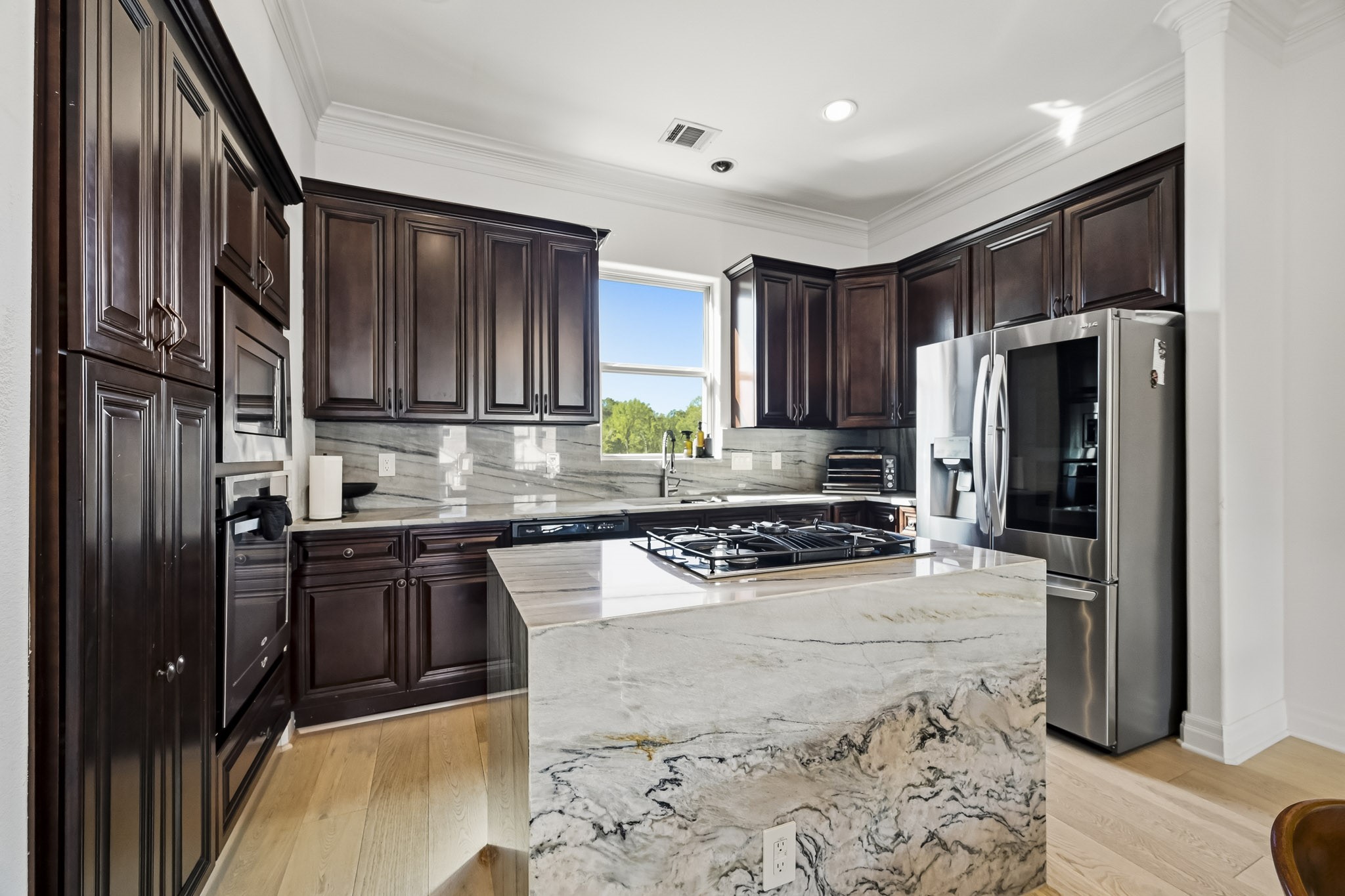 Elegant kitchen featuring rich dark wood cabinetry, a sleek stainless steel refrigerator, and a striking marble island with a built-in gas cooktop, all bathed in natural light from a well-placed window.