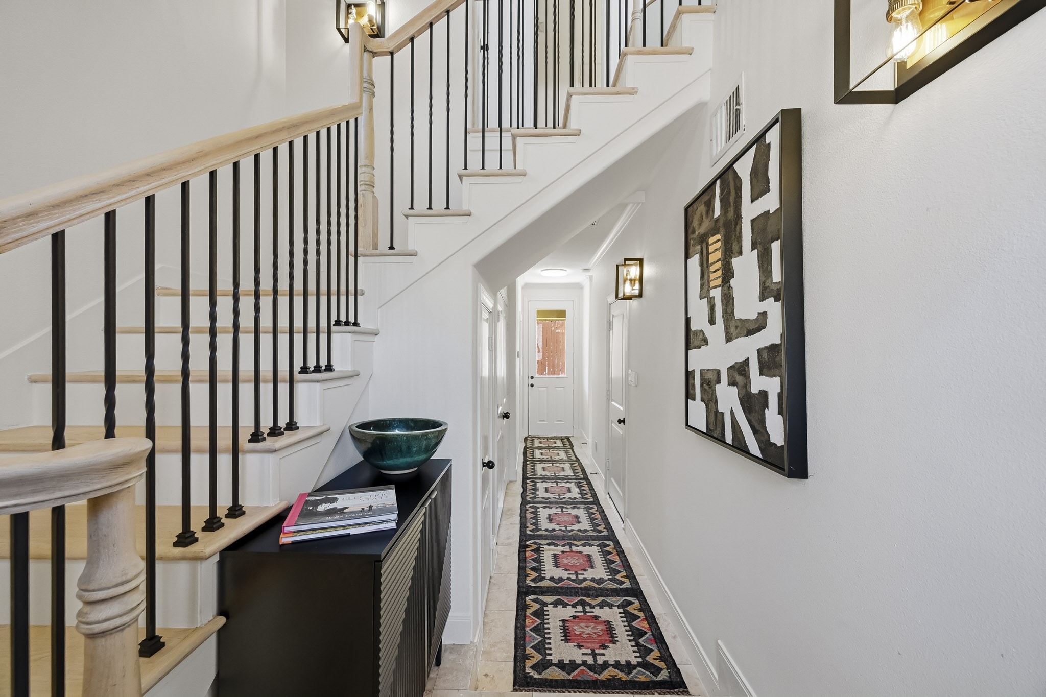 Bright and inviting entryway featuring a stylish staircase with wrought iron balusters, modern lighting, and a chic hallway runner leading to a well-lit back door.
