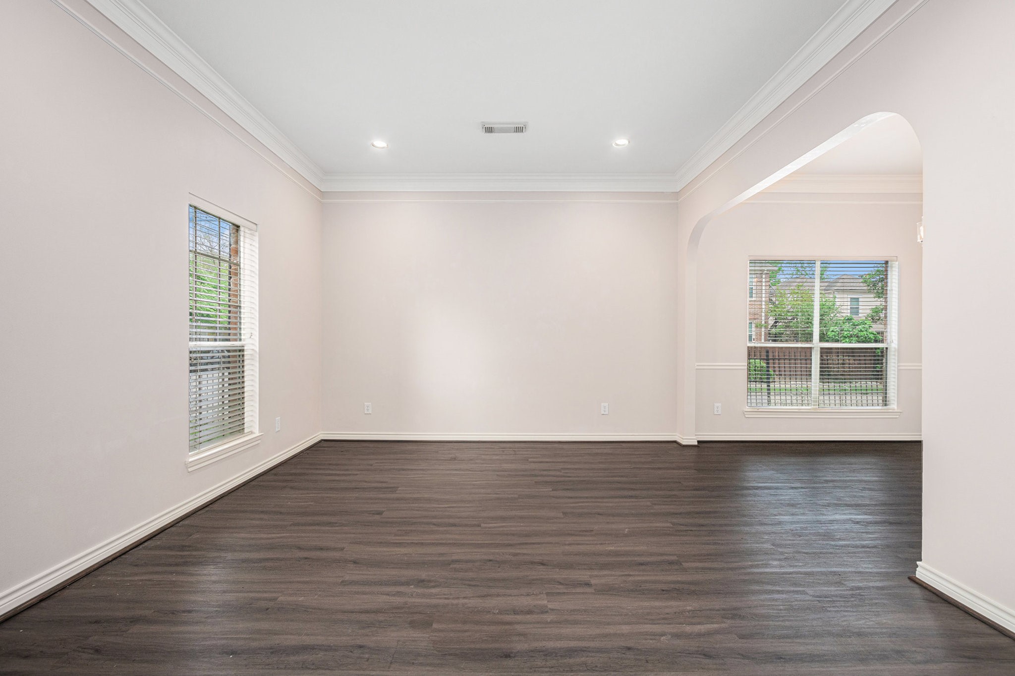 This photo showcases a bright, spacious room with light-colored walls and dark hardwood floors. It features large windows for natural light and an open layout with an arched entryway, perfect for versatile living and dining options.