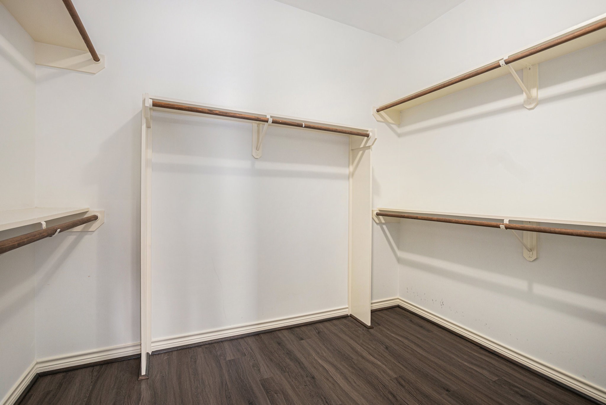 This photo shows a spacious walk-in closet with white walls, multiple wooden hanging rods, and dark wood flooring, offering ample storage space.