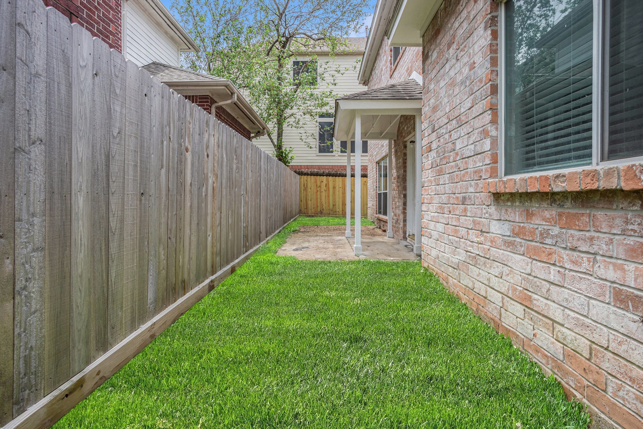 This photo shows a fenced backyard with a well-maintained grassy area, bordered by a wooden fence and a brick exterior wall of the house. There is a small covered patio area with a concrete path leading to a wooden gate.