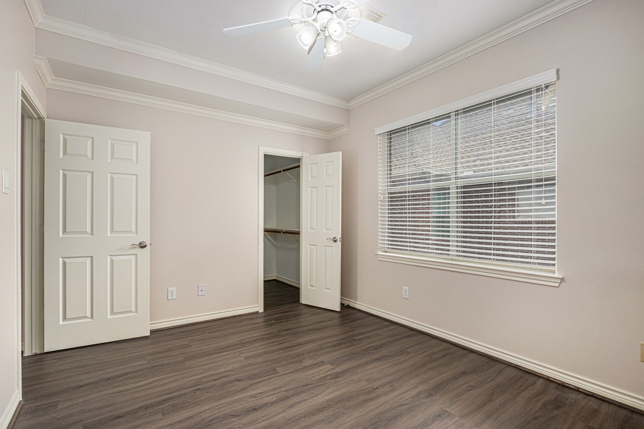 This room features sleek laminate flooring, neutral walls, and crown molding. It includes a ceiling fan, large window with blinds, and a walk-in closet.