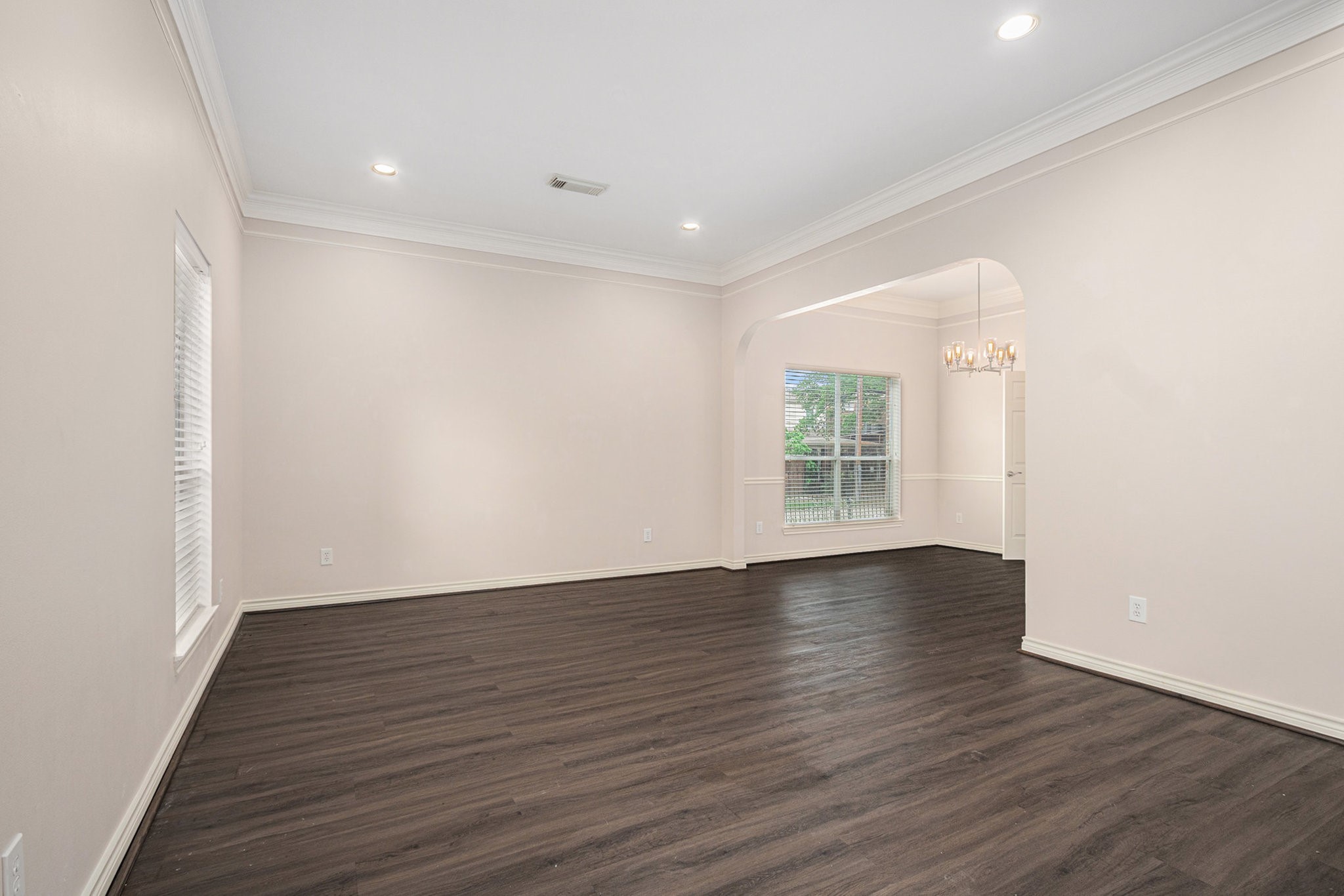 This spacious room features dark wood flooring, soft cream walls, and recessed lighting. It includes a large window for natural light and an arched opening leading to an adjacent area with a chandelier.