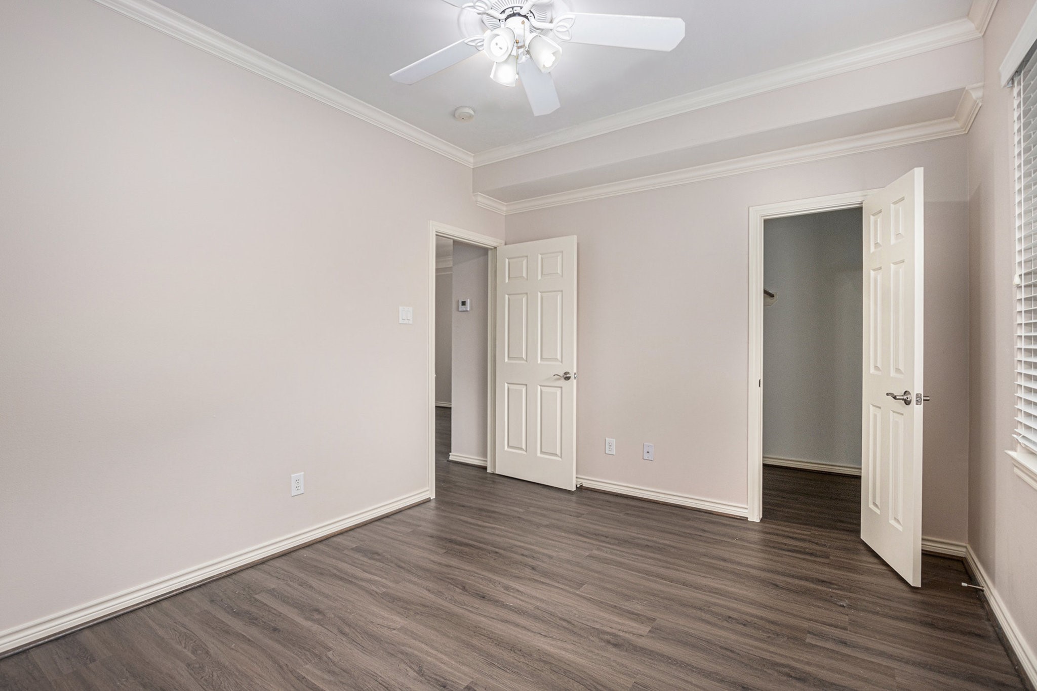 This photo shows a bright, empty room with light-colored walls and dark hardwood floors. It features a ceiling fan, crown molding, and two doorsone leading to a closet and the other to a hallway. There's a window on the right, allowing natural light to enter.