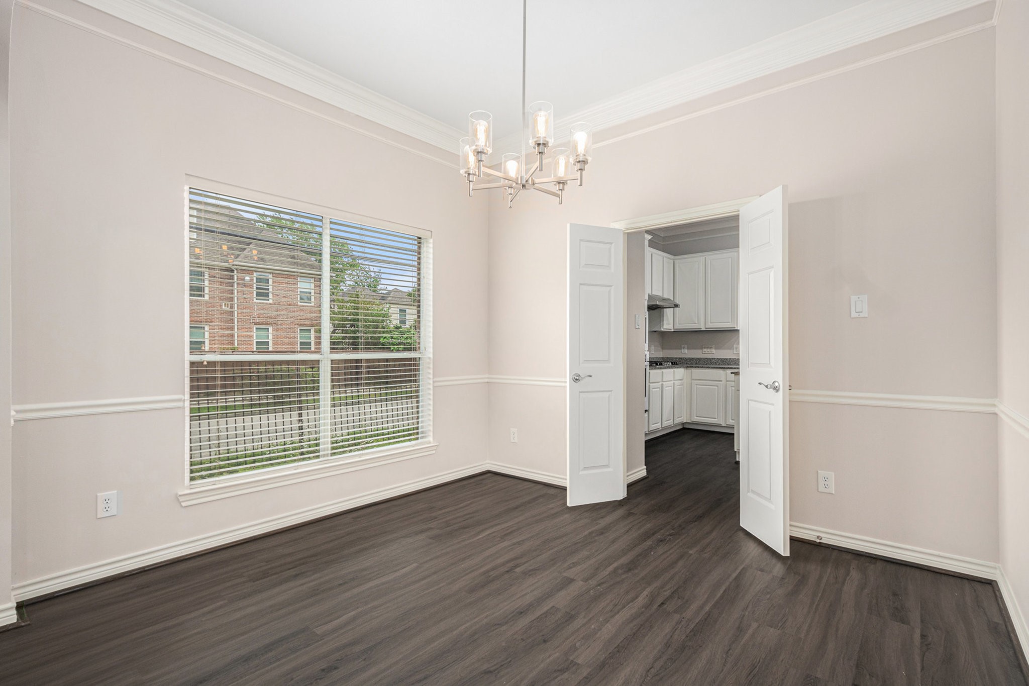 This photo shows a bright dining room with elegant crown molding, a modern chandelier, and dark wood flooring. Large windows provide natural light, and double doors lead into a white kitchen.