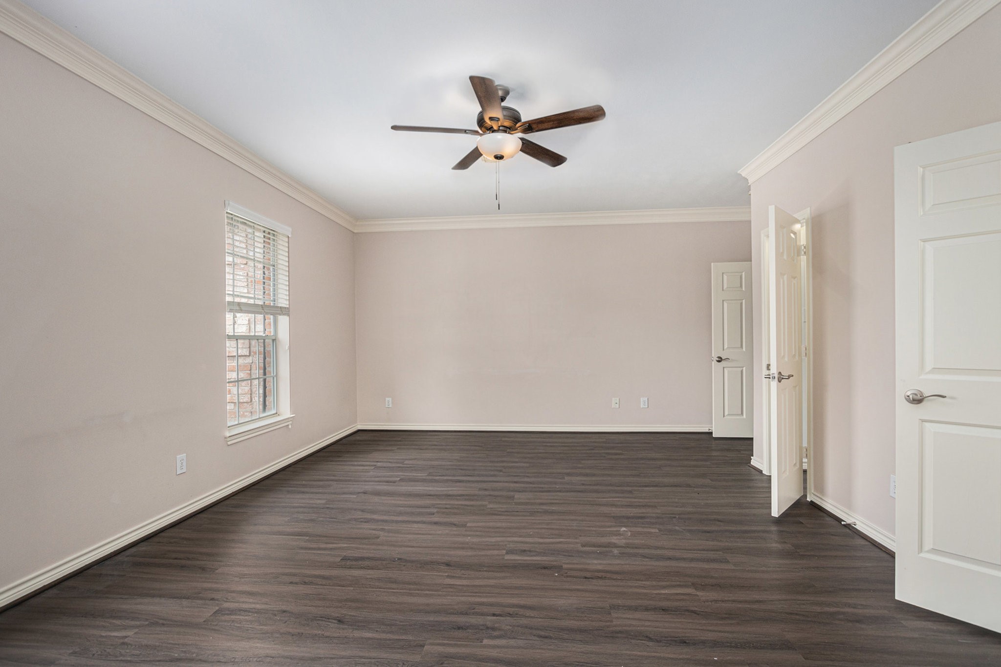 This spacious room features neutral walls, dark wood flooring, a ceiling fan, and ample natural light from a large window, creating a bright and inviting atmosphere.