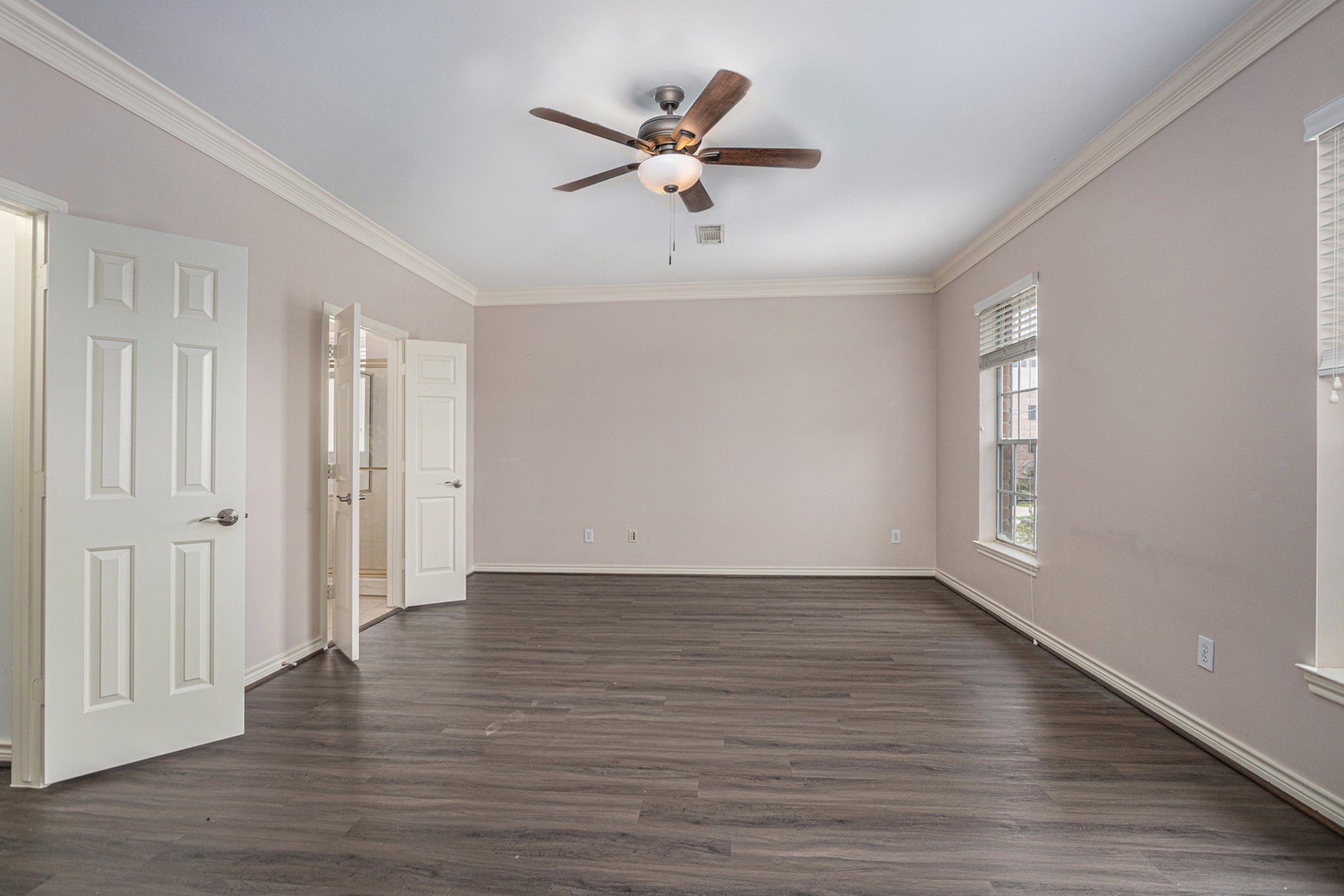 This spacious room features elegant crown molding, a ceiling fan, and rich wood flooring. It offers plenty of natural light through large windows, and has a neutral color palette, creating a welcoming atmosphere.