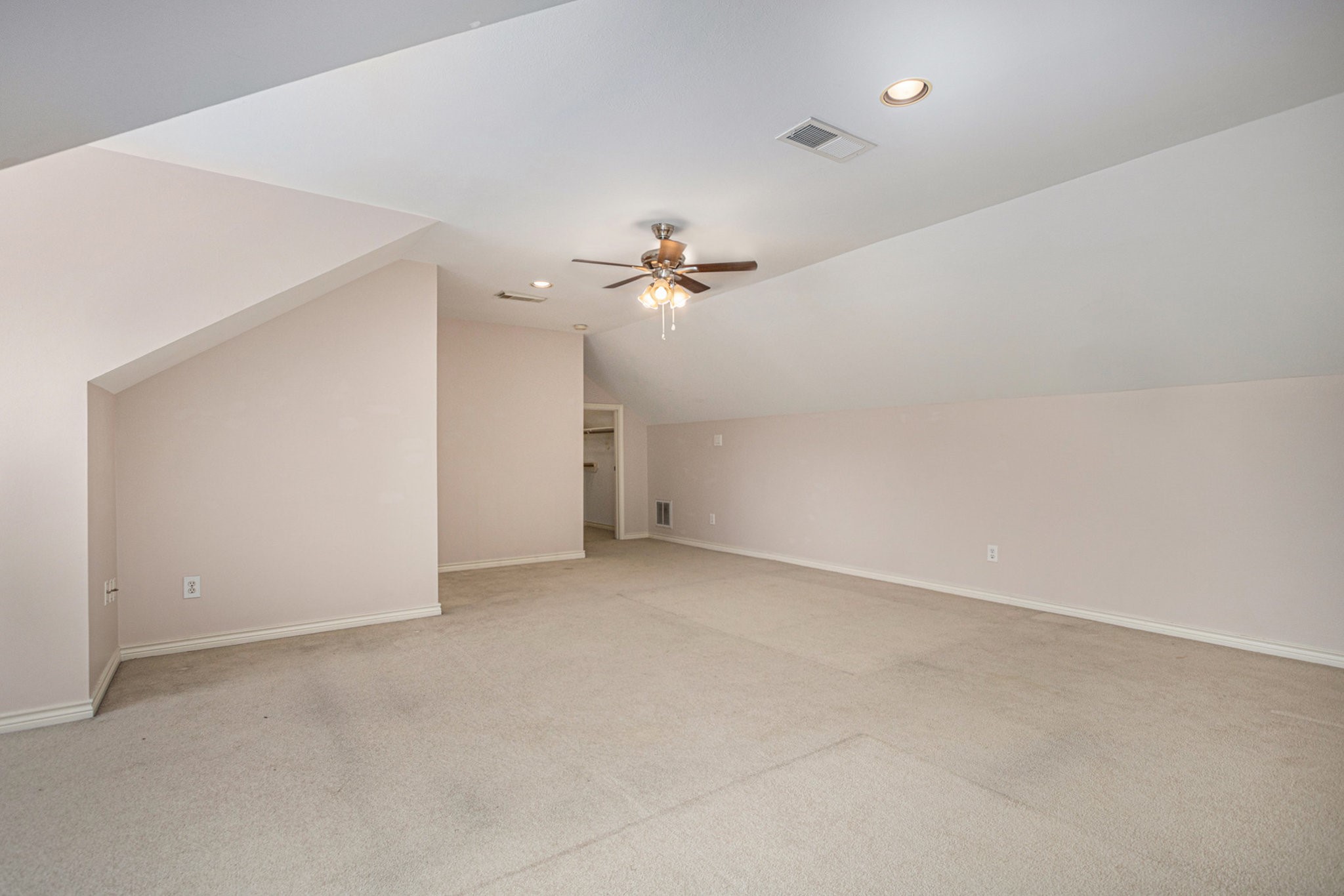 This spacious, neutral-toned attic room features a sloped ceiling, recessed lighting, and a ceiling fan, making it versatile for various uses.