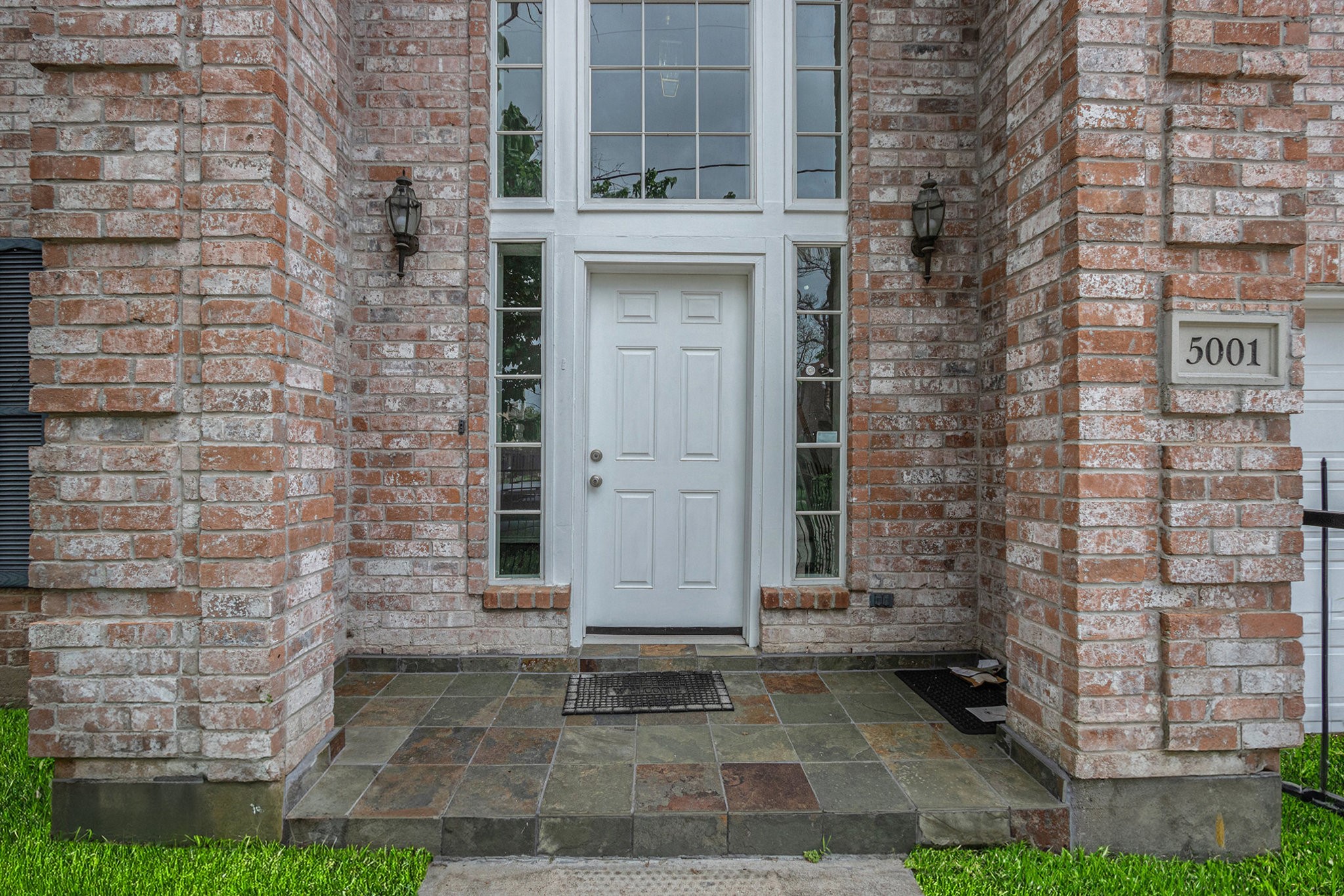 This entrance features a brick facade with a welcoming white door and large windows, allowing natural light into the home. The slate-tiled porch adds a touch of elegance, complemented by decorative outdoor lighting.