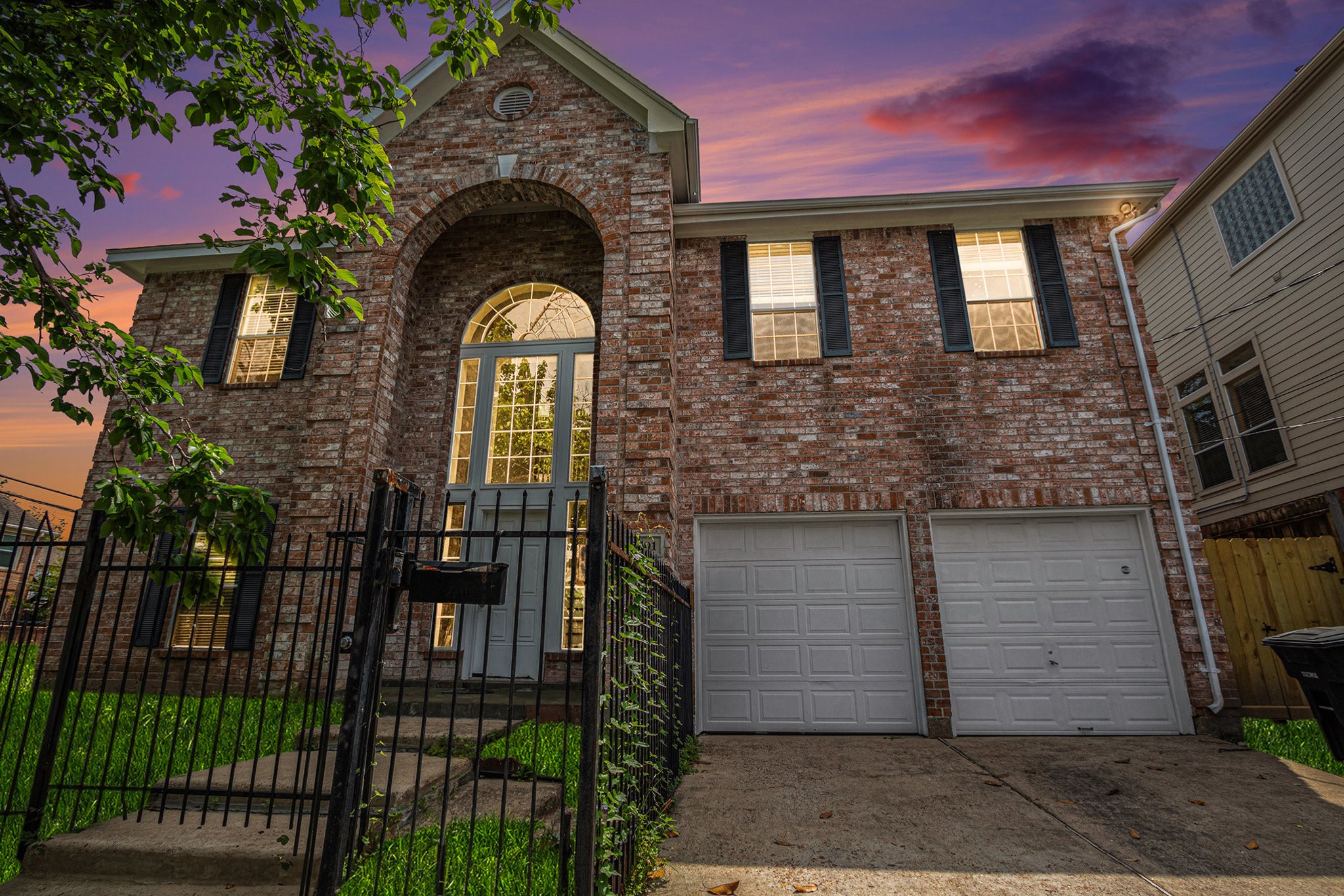 This photo features a two-story brick home with an elegant arched entryway and large windows. It has a double garage and is surrounded by a neatly fenced yard. The sunset sky adds a warm, inviting glow to the property.