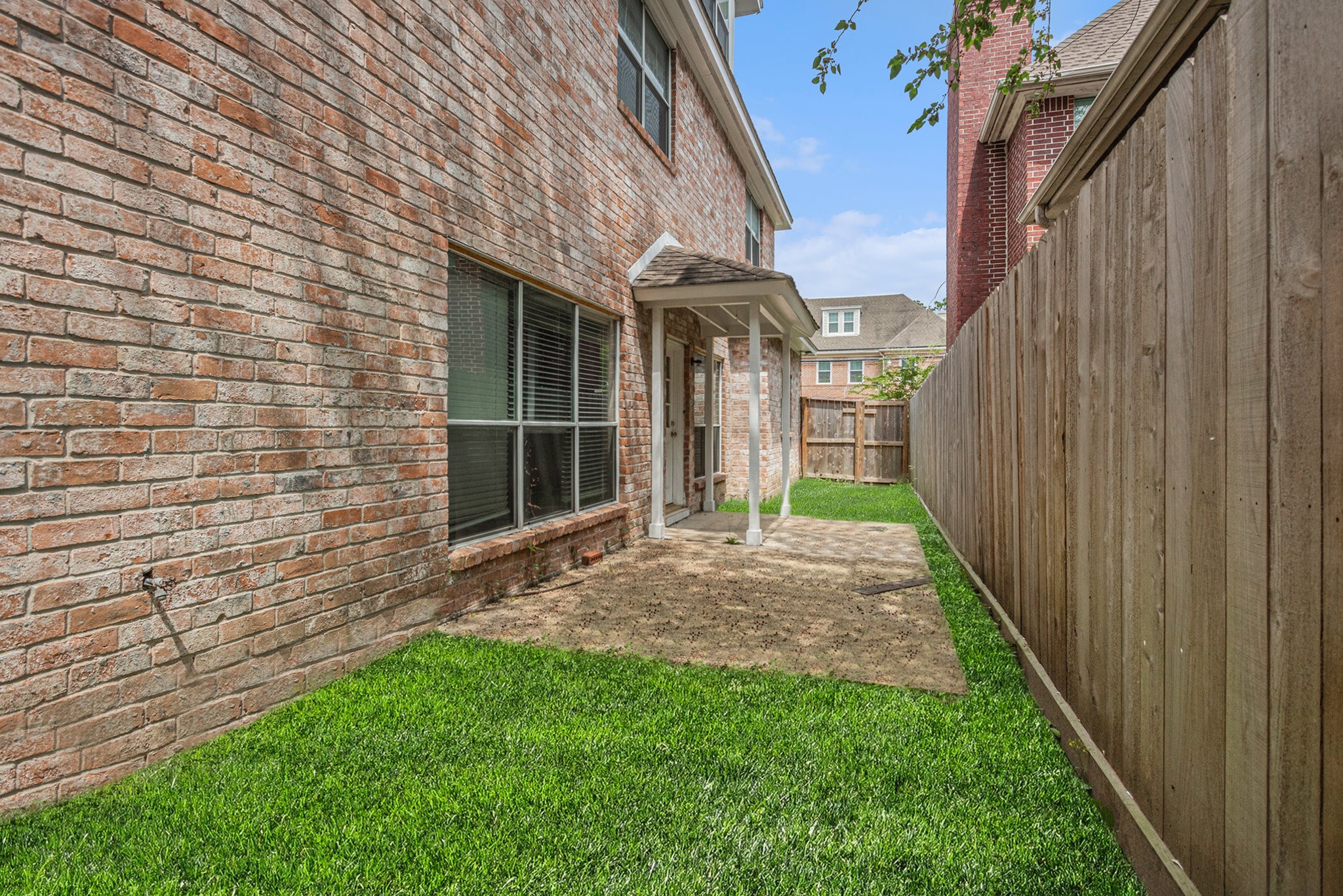 This photo shows a fenced backyard with a brick exterior, lush green grass, a small covered patio, and a wooden fence for privacy. Ideal for low-maintenance outdoor space.