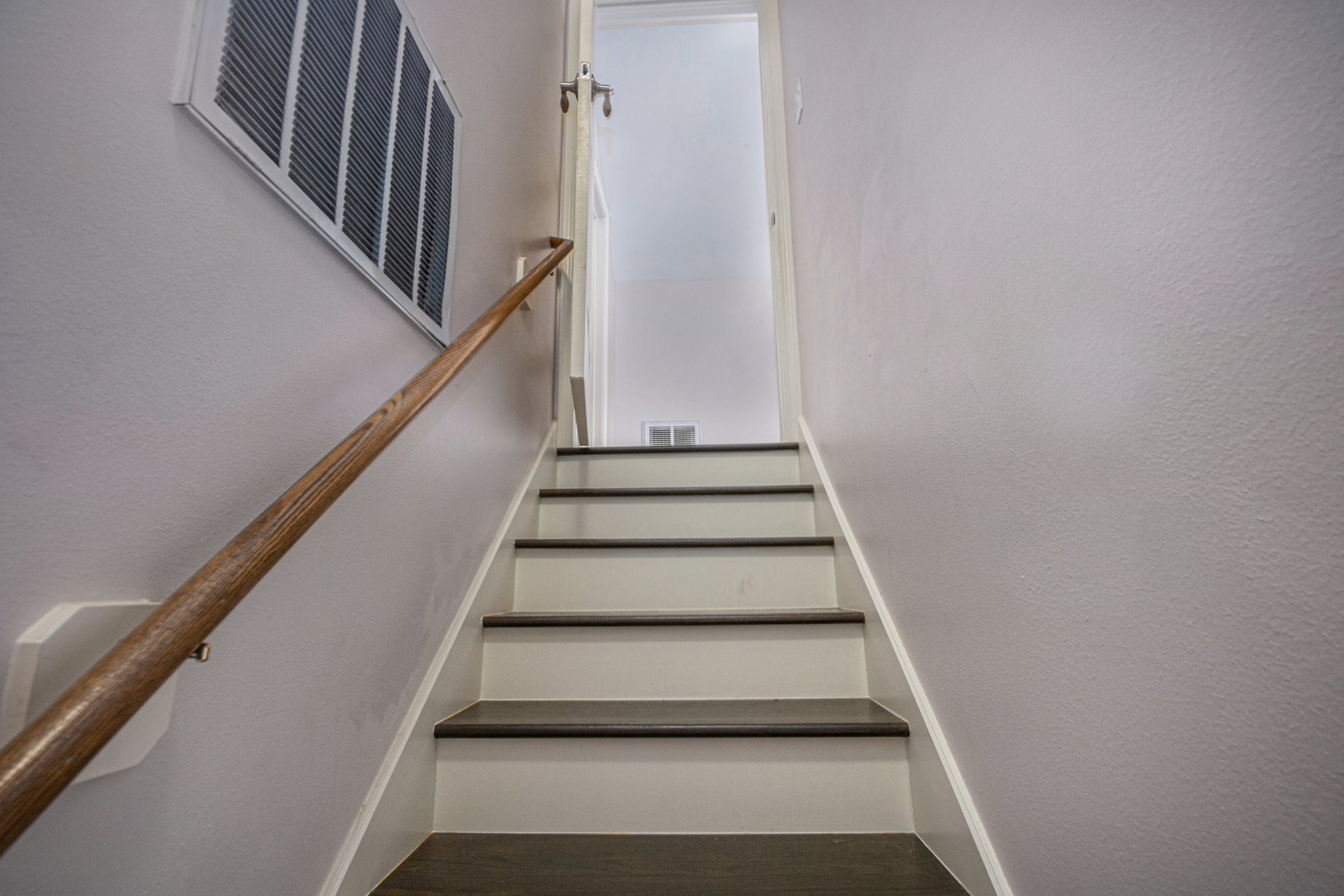 This photo shows a staircase with dark wood steps and white risers, leading up to a doorway. The walls are painted a light neutral color, and a wooden handrail lines the left side. An air vent is visible on the left wall.