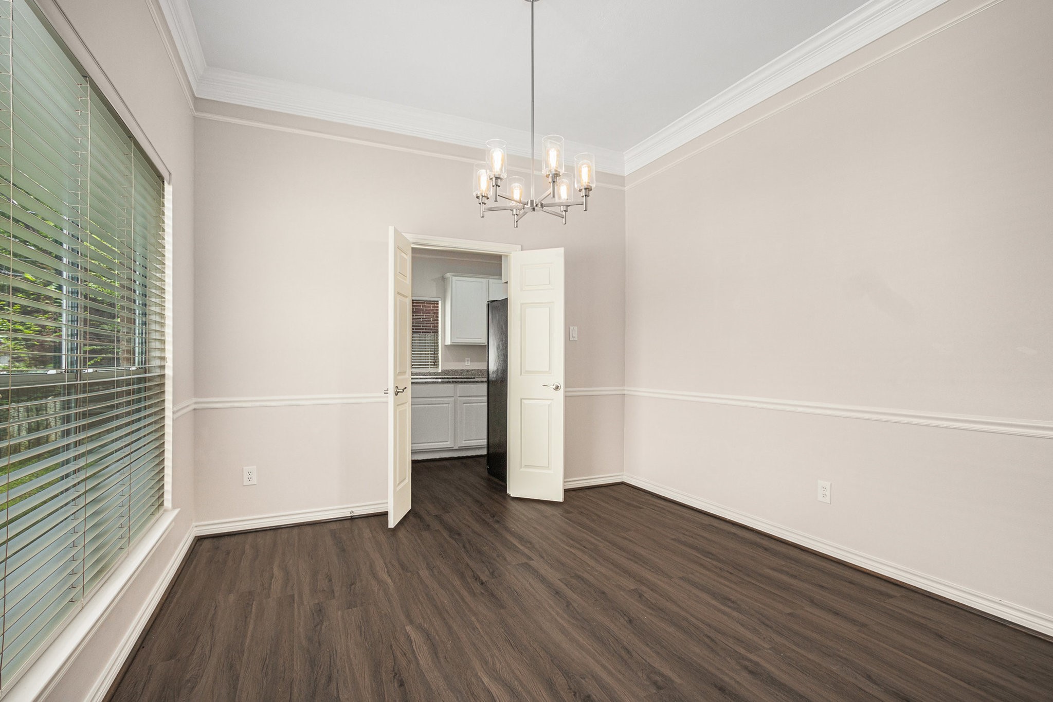 This room features elegant wood flooring, a large window with blinds for natural light, and a modern chandelier. It opens into a kitchen area through double doors, offering a seamless flow between spaces. Perfect for dining or as a versatile living area.