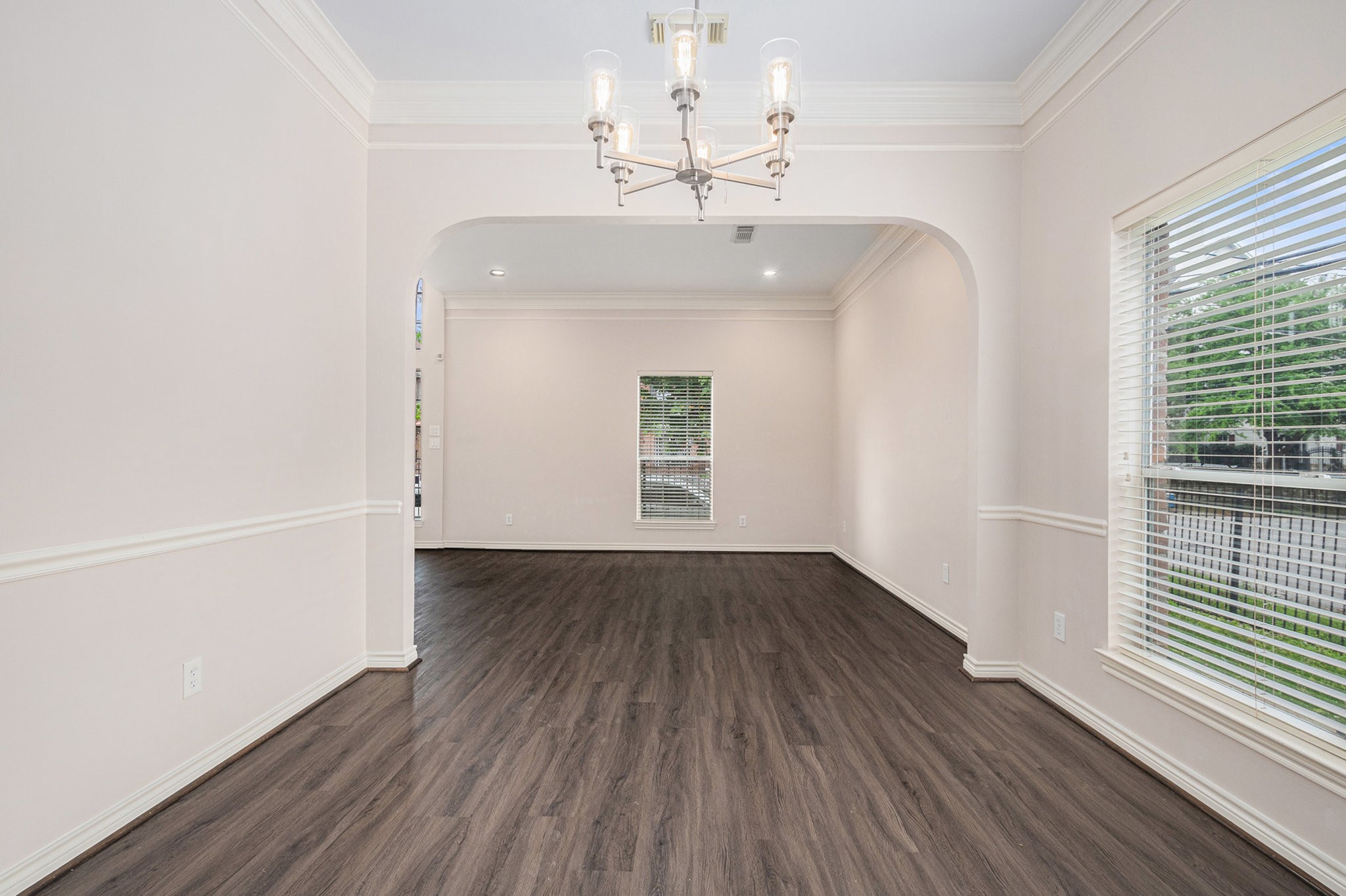 Bright, spacious room with elegant wood flooring, large windows for natural light, and a modern chandelier. Ideal for dining or living space.