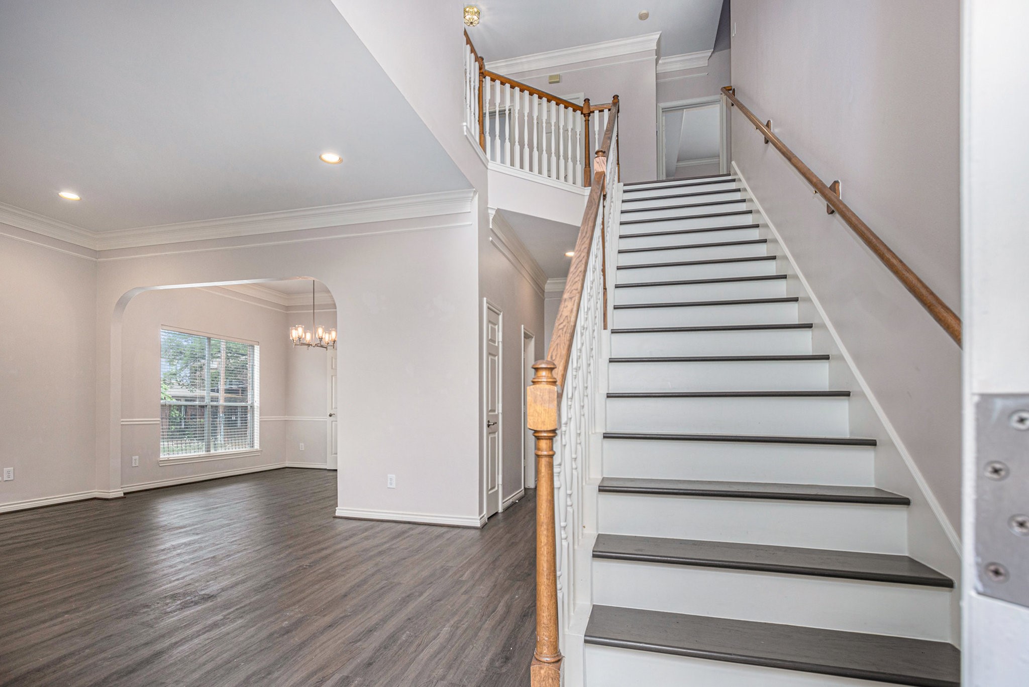 This photo showcases a bright, open entryway with elegant hardwood flooring, a staircase with wooden handrails, and a view into a spacious dining area featuring large windows and a modern chandelier.