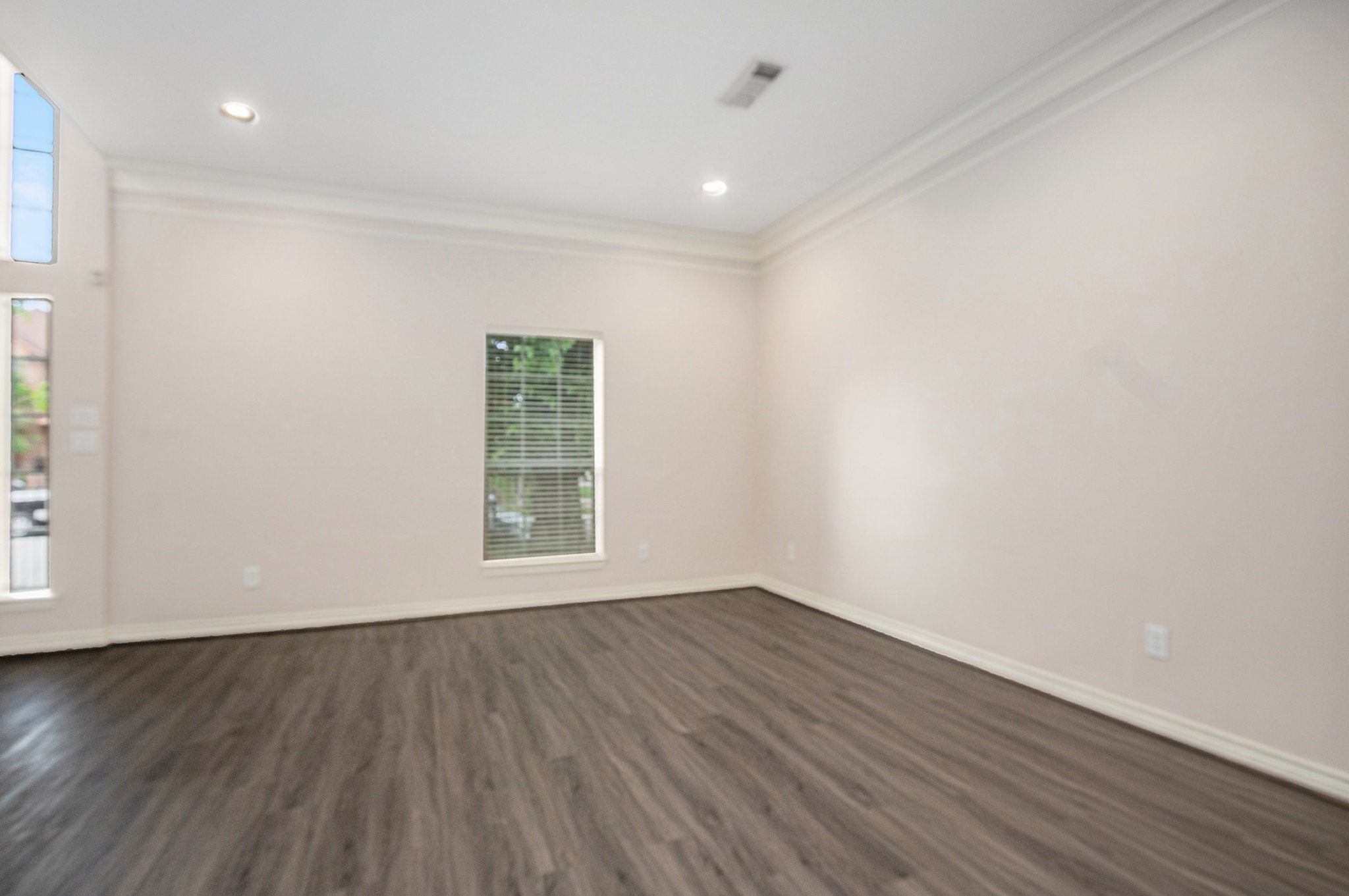 This room features neutral walls, a single window with blinds, recessed lighting, and dark wood flooring, creating a bright and modern space.