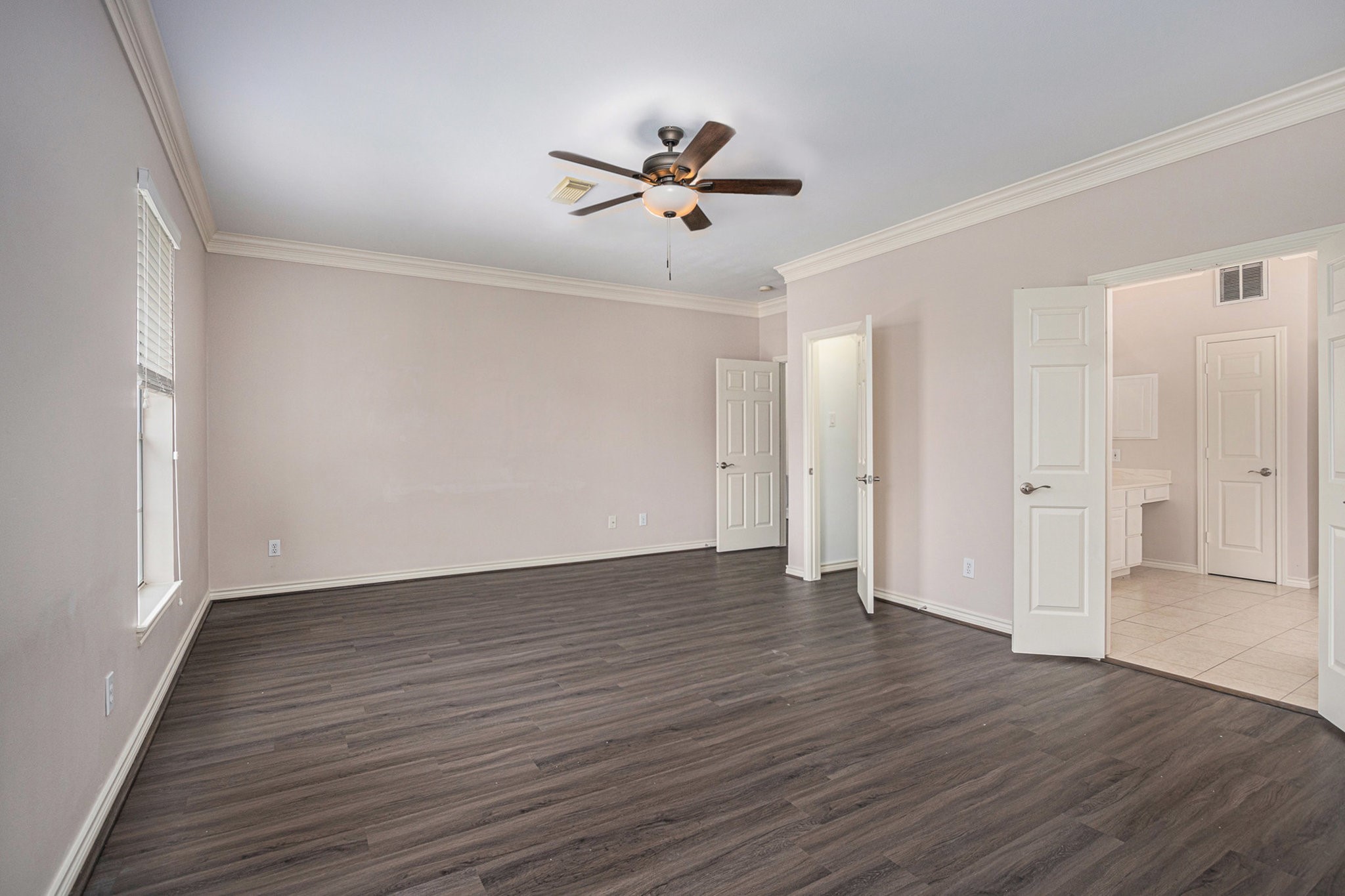 Spacious bedroom with dark wood flooring, neutral walls, and a ceiling fan. It features two doors leading to a bathroom and a closet, offering ample natural light through large windows.