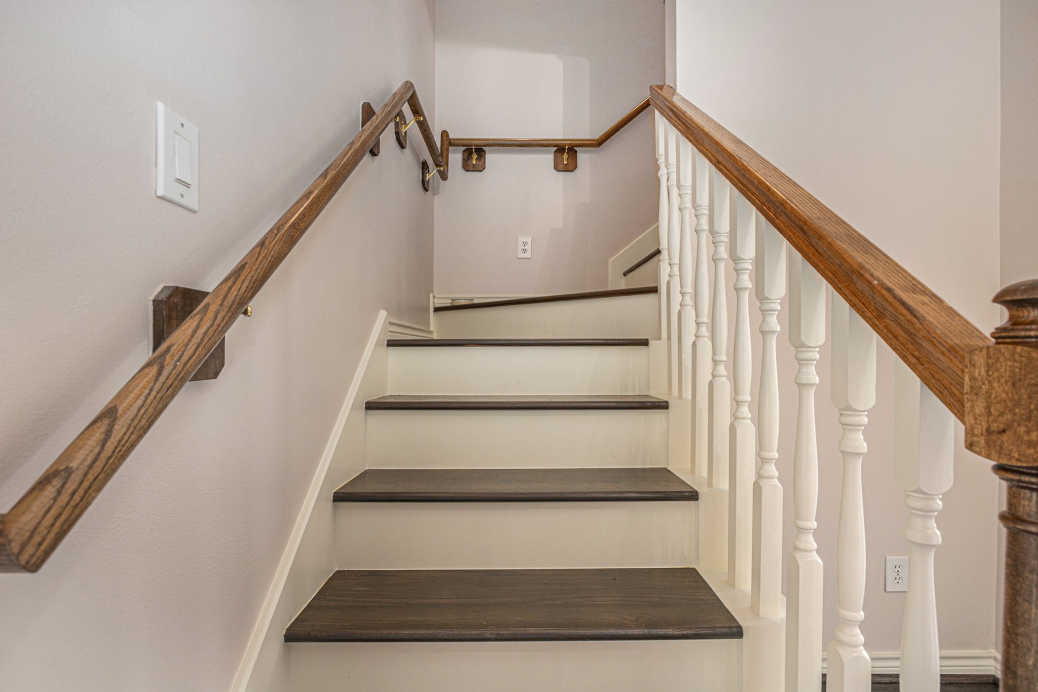 This photo showcases a staircase with dark wooden steps and white spindles, complemented by wooden handrails on both sides, leading to a landing. The walls are painted in a light, neutral color.