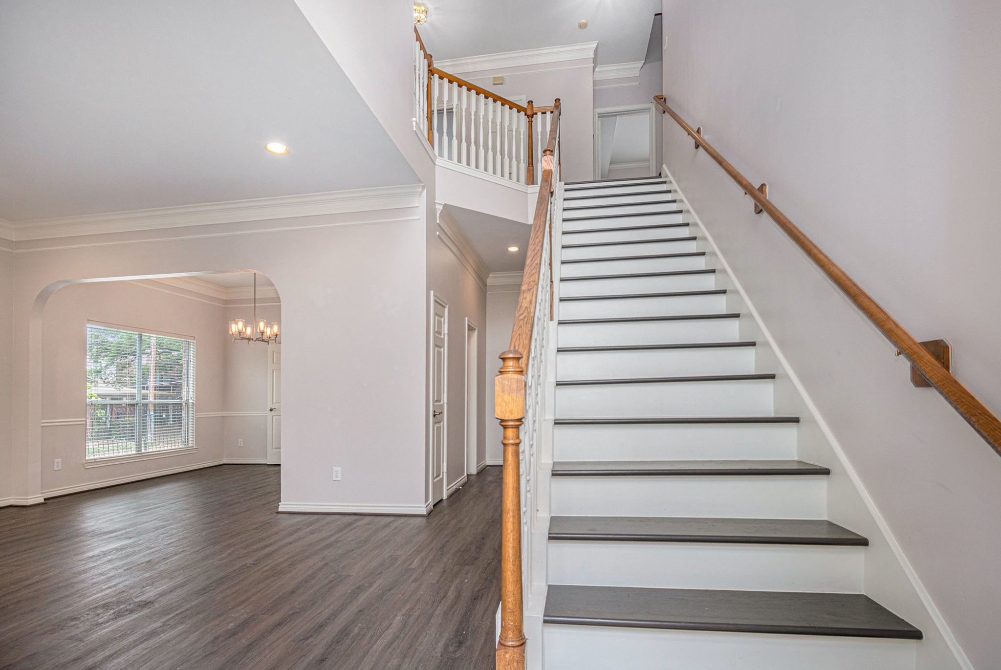 This photo showcases a bright, spacious entryway with elegant hardwood flooring, a prominent staircase with a wooden banister, and an open view into a dining area featuring a large window and chandelier.