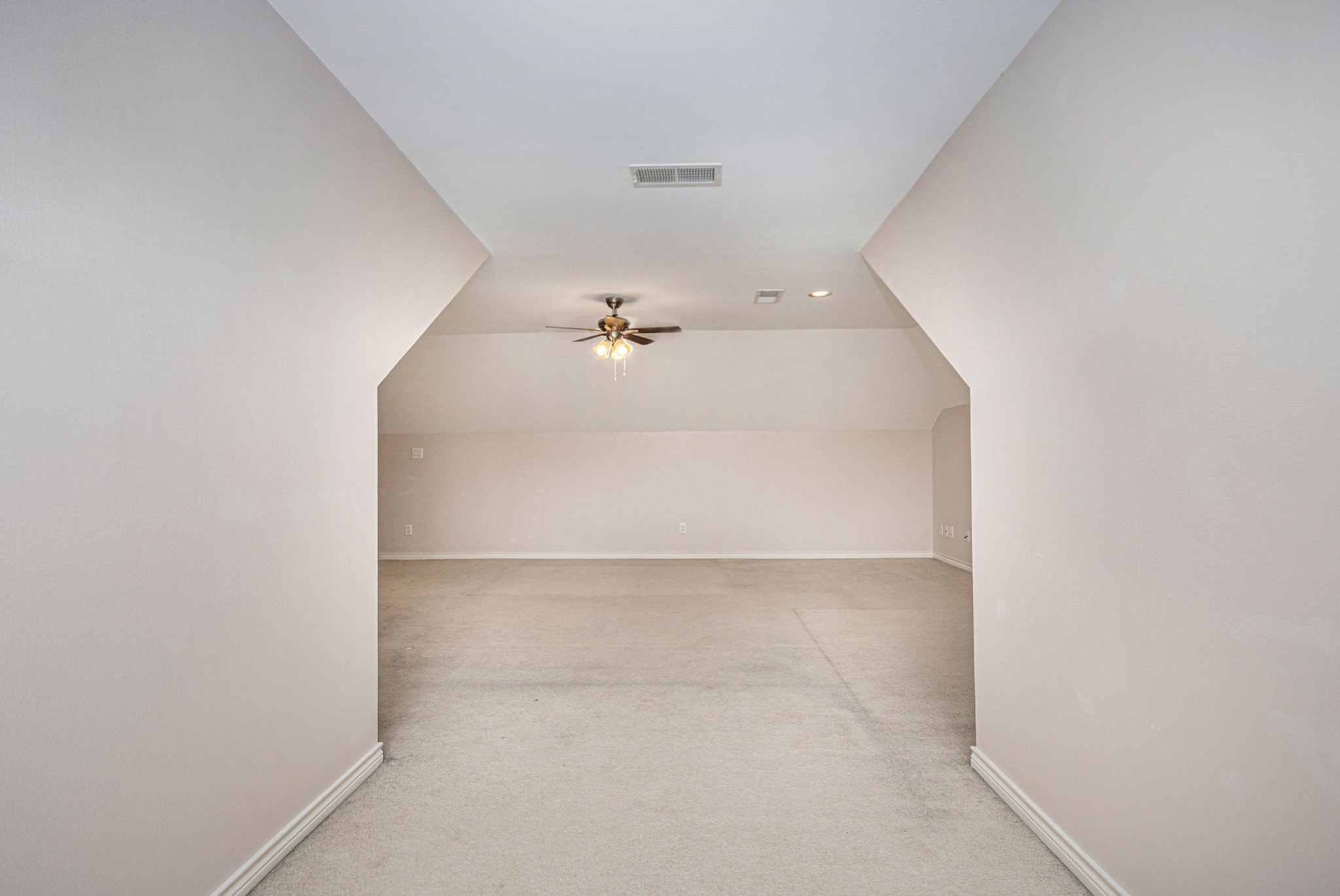 Spacious attic room with angled ceilings, neutral walls, and carpeted flooring, featuring a ceiling fan and recessed lighting. Ideal for a versatile living space or storage.