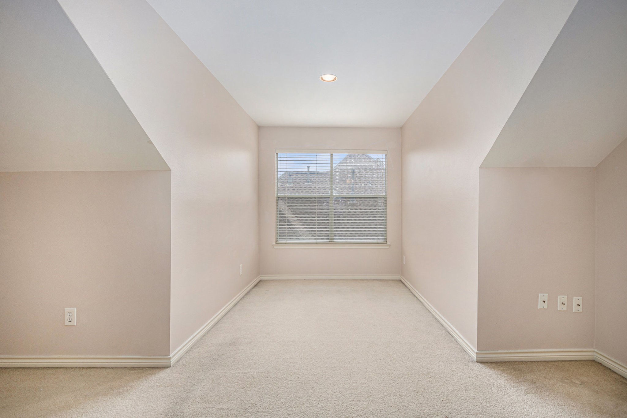 This is a cozy, carpeted attic space with sloped ceilings and a window providing natural light. Ideal for a small office or reading nook.