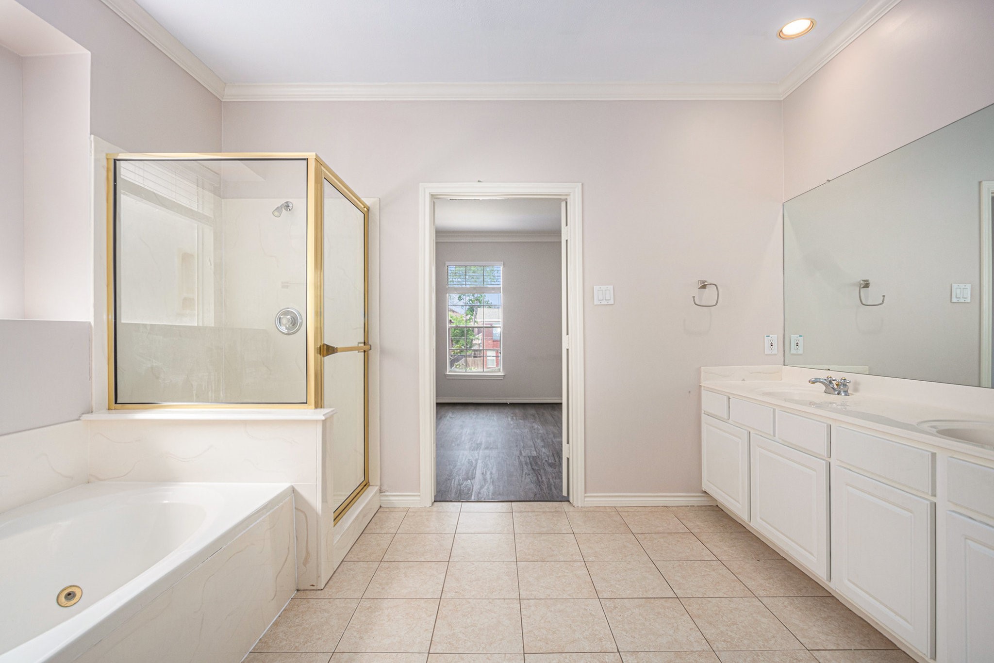 This photo showcases a spacious bathroom with a large soaking tub, glass-enclosed shower, dual-sink vanity, and neutral tile flooring. A doorway leads to an adjacent room with a window.
