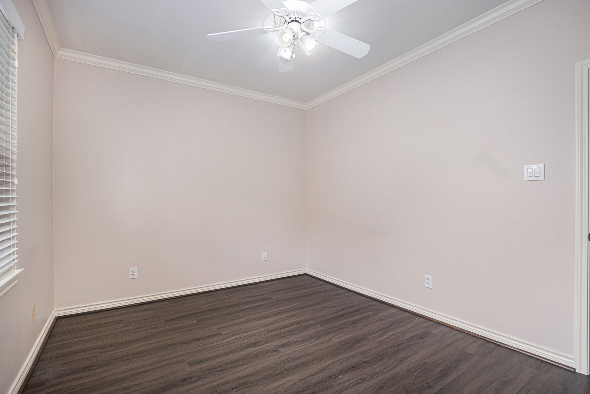This room features neutral walls, dark wood flooring, a ceiling fan with lights, and a window with blinds, offering a bright and versatile space.