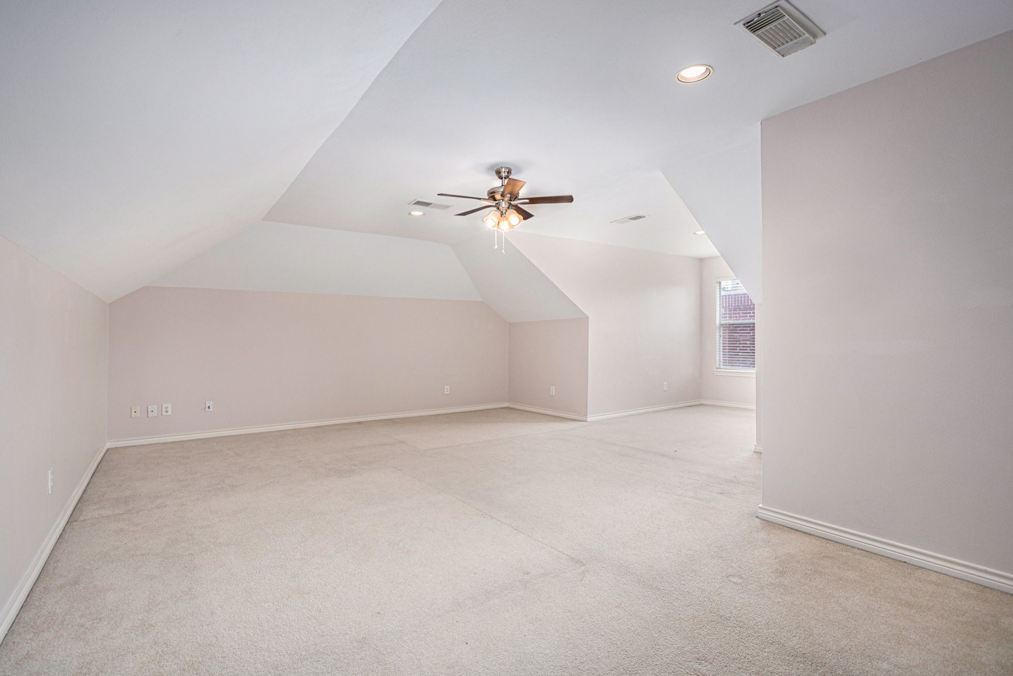 This spacious, light-filled room features a sloped ceiling and neutral carpet, perfect for a bedroom or home office. It includes a ceiling fan and a window for natural light.