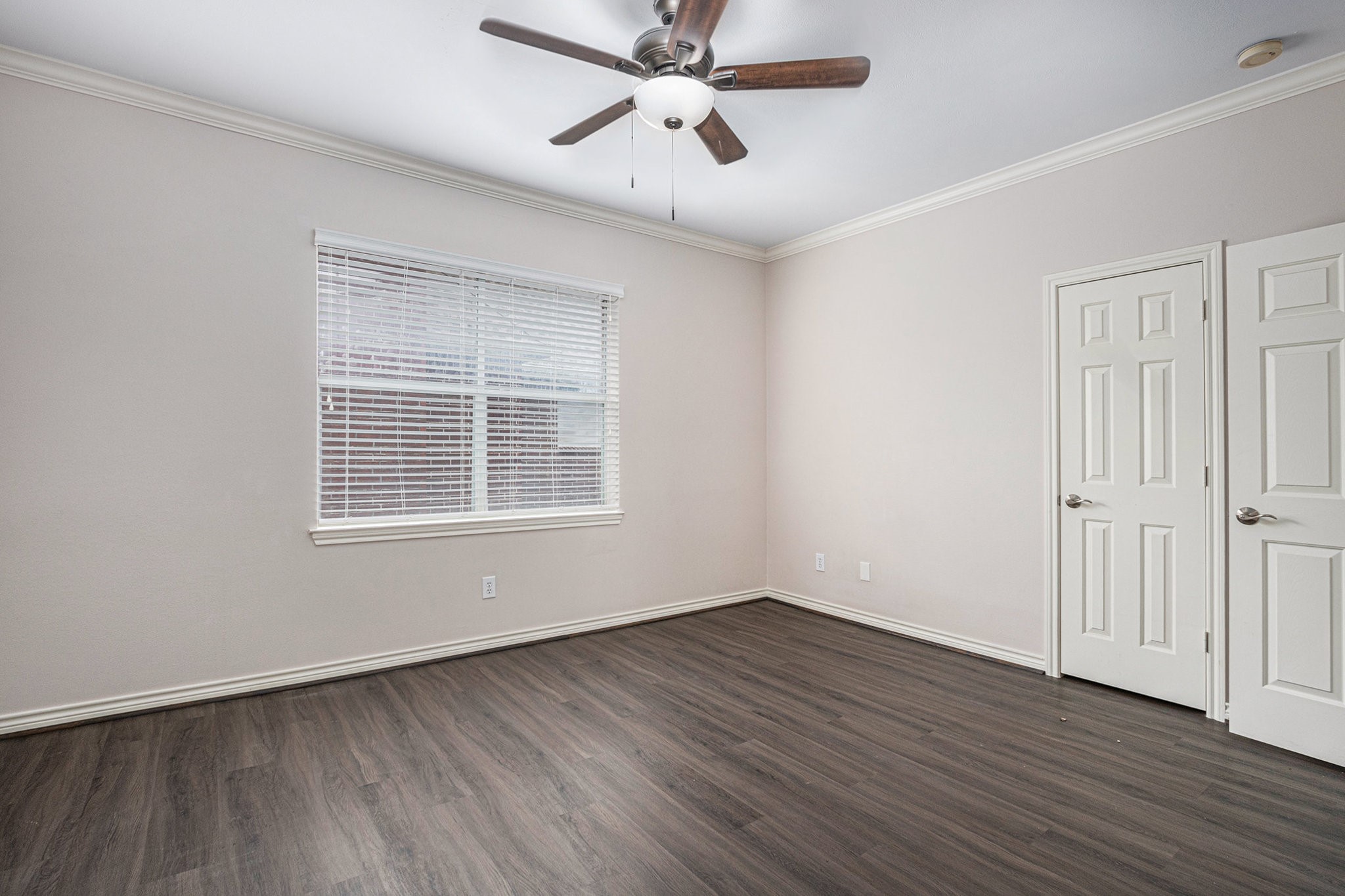 This room features laminate wood flooring, neutral walls, and a ceiling fan with light. A large window provides natural light, and there are two white doors, likely for closet space.