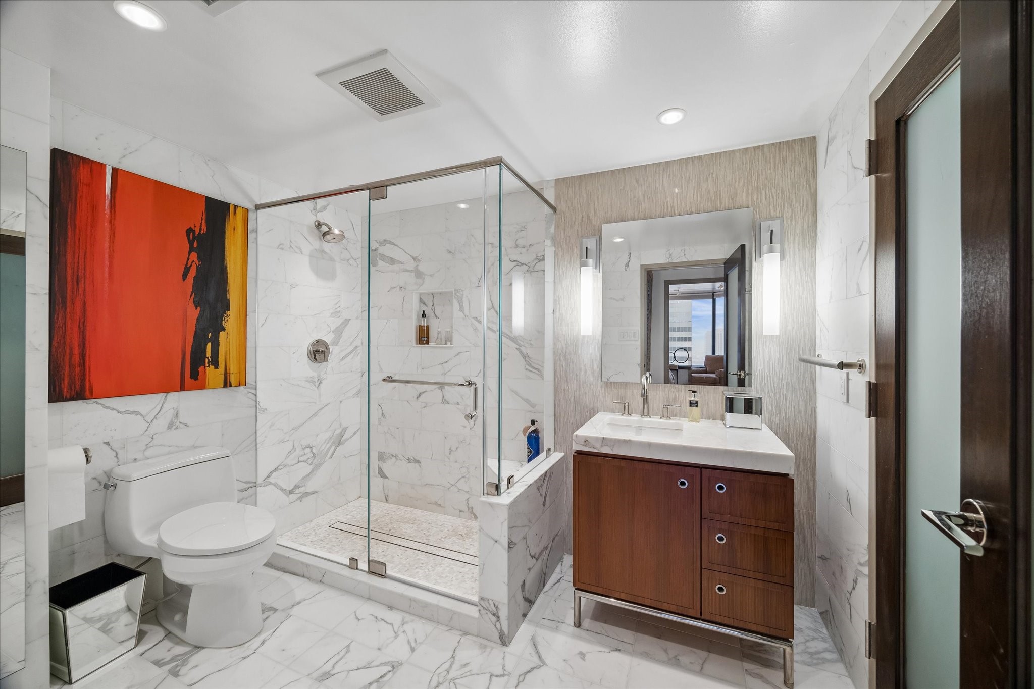 Glass enclosed shower, mosaic shower floor and veined tile define the bath. Vanity drawers and cabinets provide storage beside the sink, with vertical lighting flanking the mirror.
