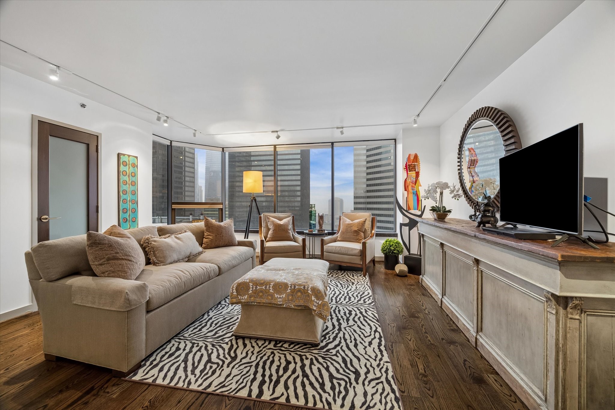 Expansive windows frame downtown views across the living room. Automated Lutron shades provide convenient control over natural light, while wood floors and track lighting define the open space.