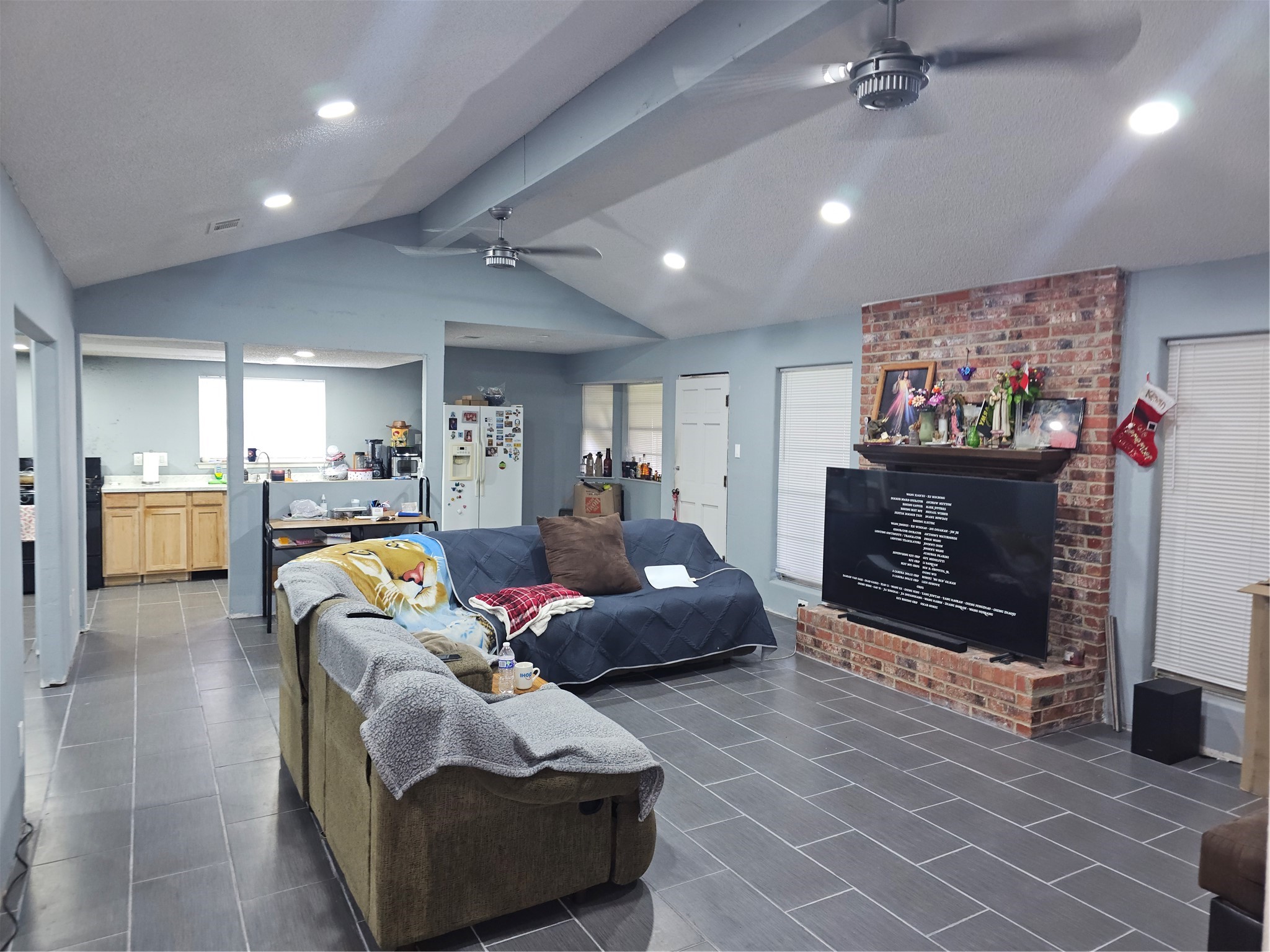 Open-concept living area features vaulted ceilings, a classic brick fireplace, tile flooring, and abundant natural light.