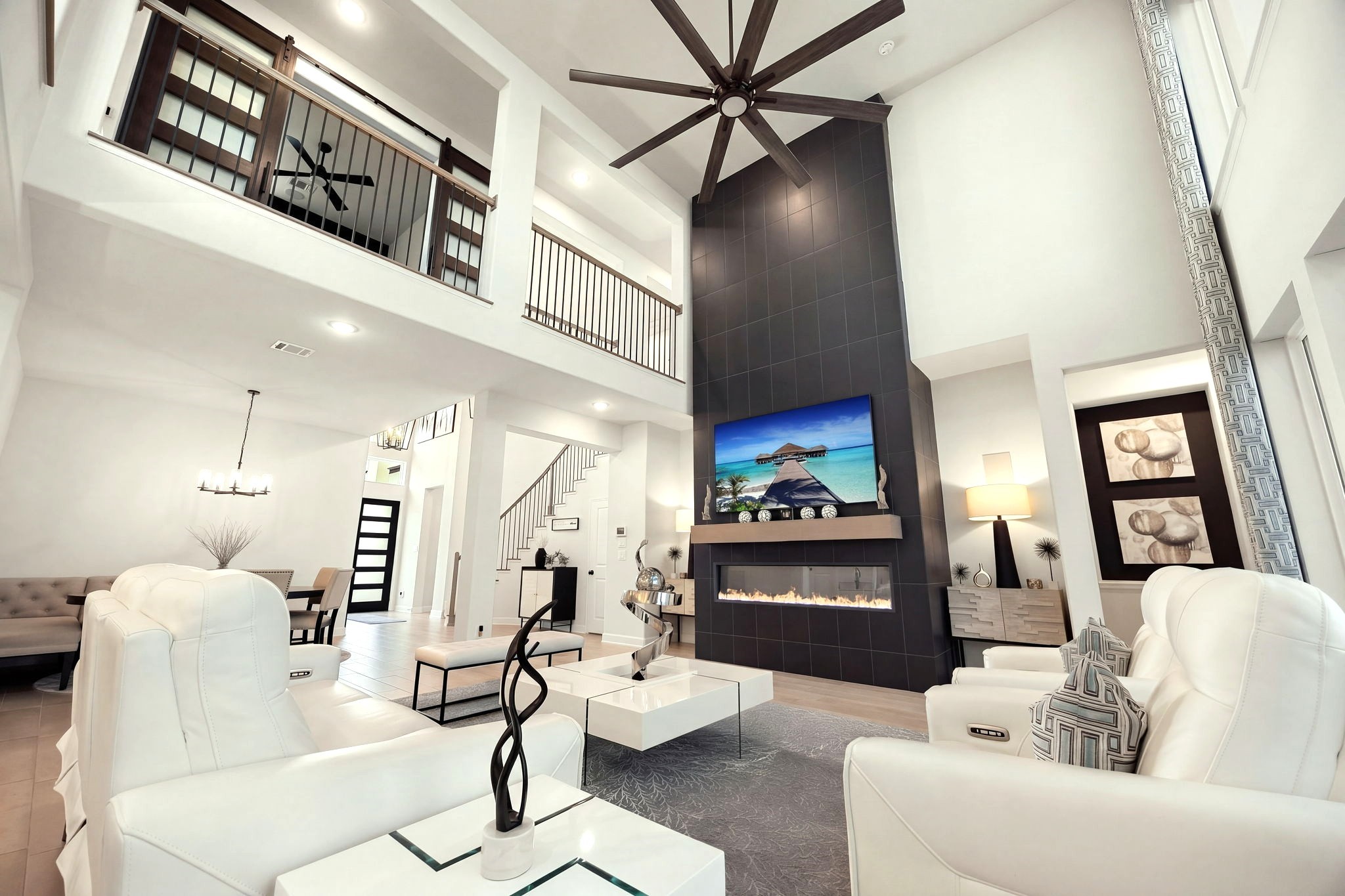 A dramatic upward view highlights the soaring ceilings, oversized upgraded black ceiling fan, and striking floor-to-ceiling stone fireplace, complemented by elegant upstairs iron railings that enhance the home's impressive architectural design.