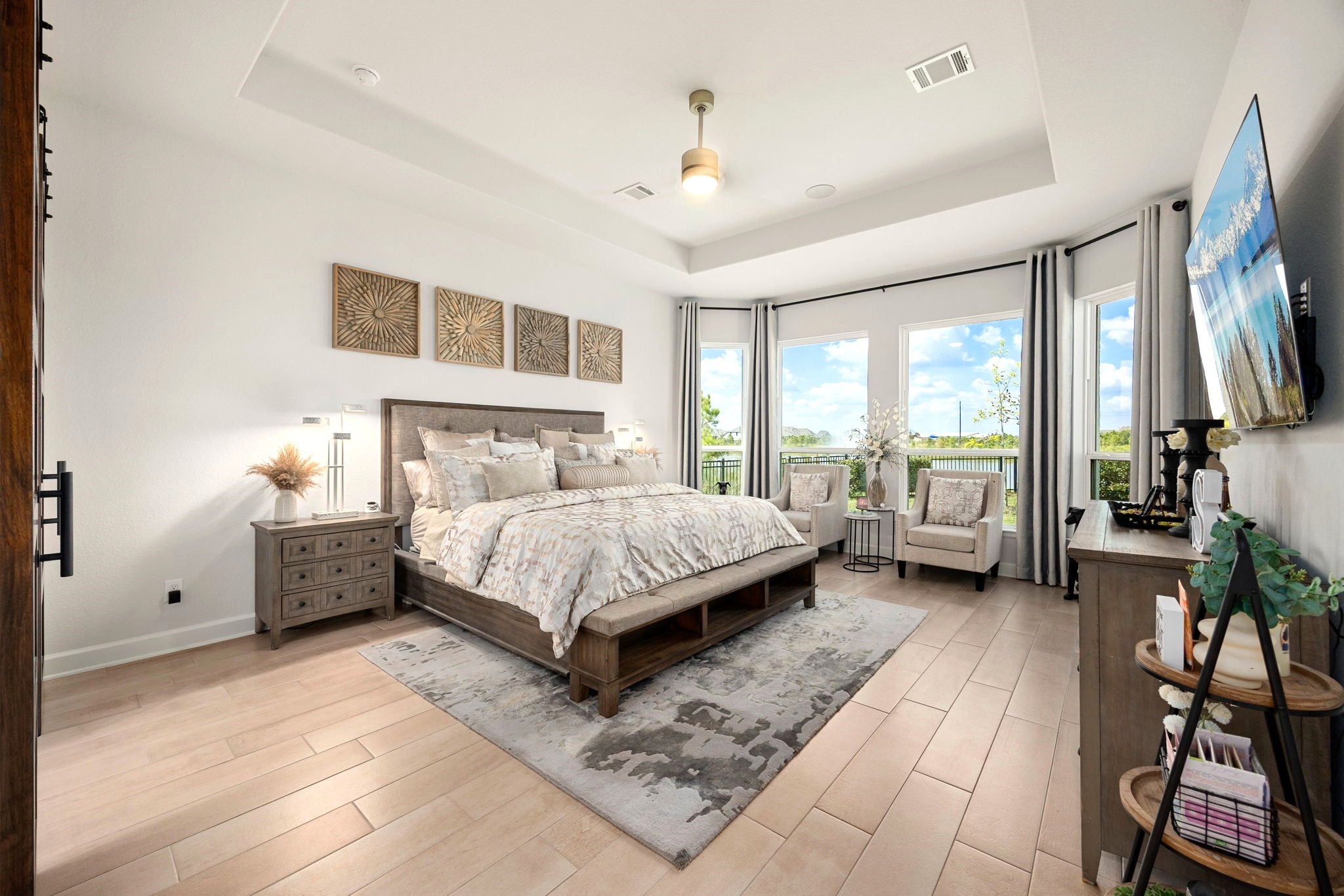 An extended primary suite features upgraded wood tile flooring, an elegant elevated tray ceiling, and four expansive windows framing serene lake views, creating a spacious and luxurious private retreat filled with natural light.
