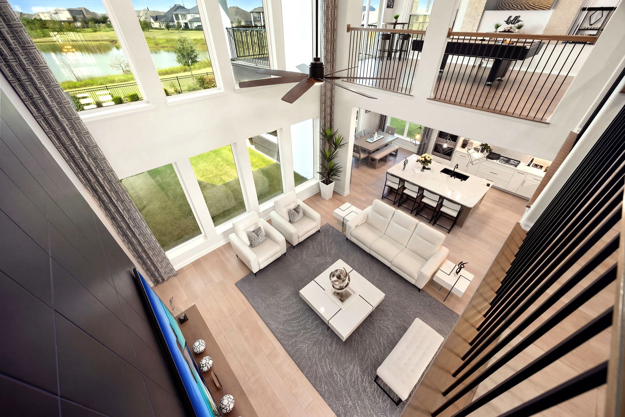 An impressive second-floor vantage point overlooks the grand living room below, showcasing elegant iron railing details, soaring ceilings, and expansive windows framing beautiful lake views for a breathtaking blend of architectural elegance and serene luxury.