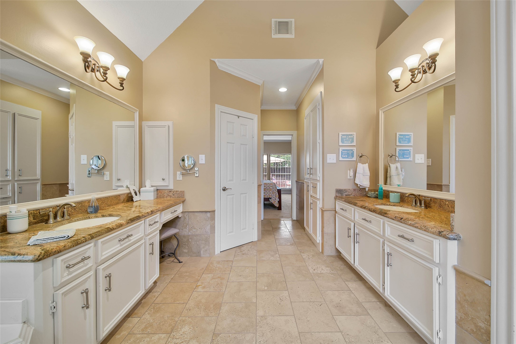 This spacious bathroom features dual vanities with granite countertops, elegant lighting fixtures, and ample cabinet storage. The tile flooring complements the neutral wall tones, creating a warm and inviting atmosphere. A doorway leads to the adjacent bedroom.
