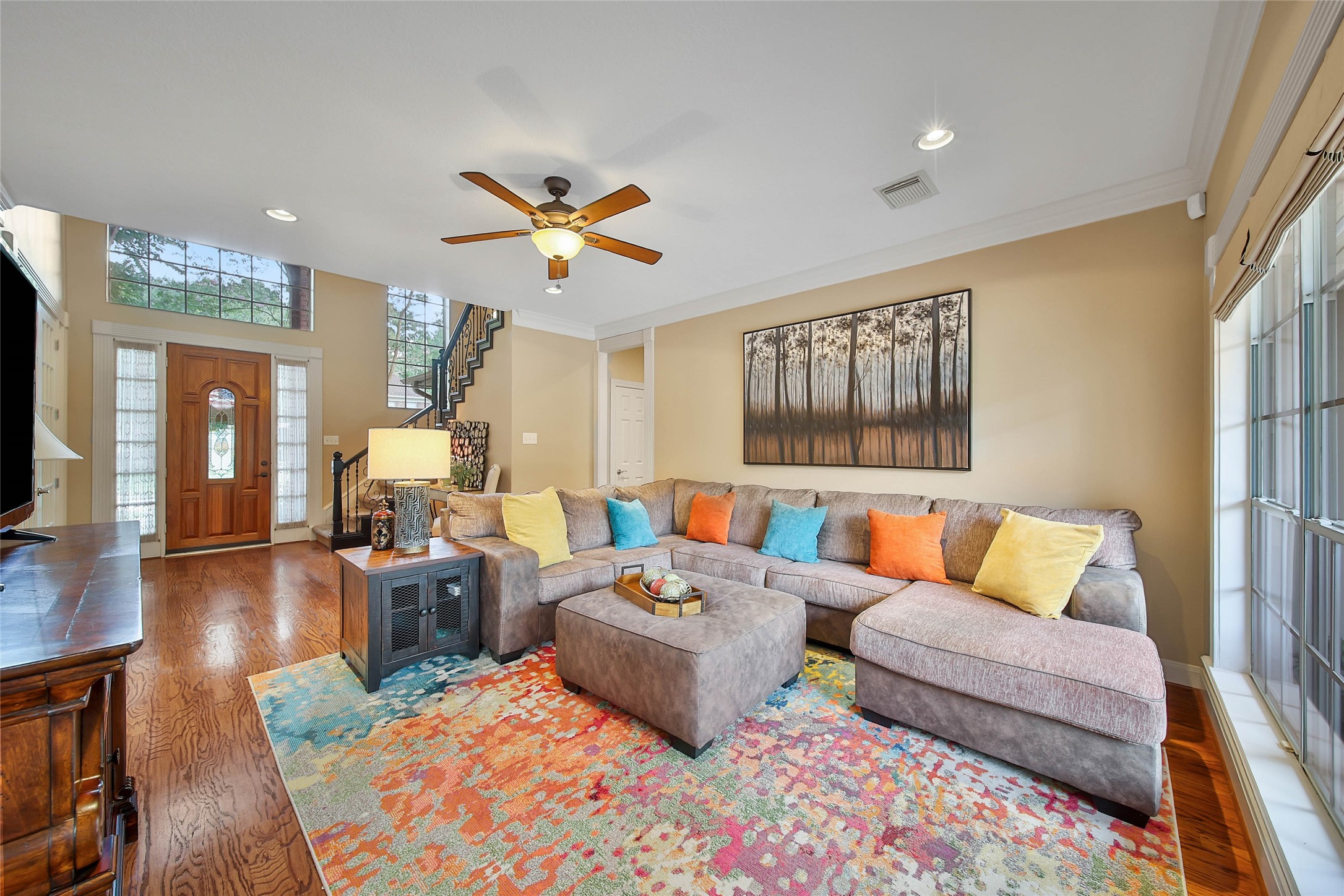 Bright and welcoming living room featuring a large sectional sofa with colorful cushions, a vibrant area rug, and hardwood floors. The space is enhanced by a ceiling fan, large windows, and a view of the staircase and wooden front door, creating an inviting atmosphere.