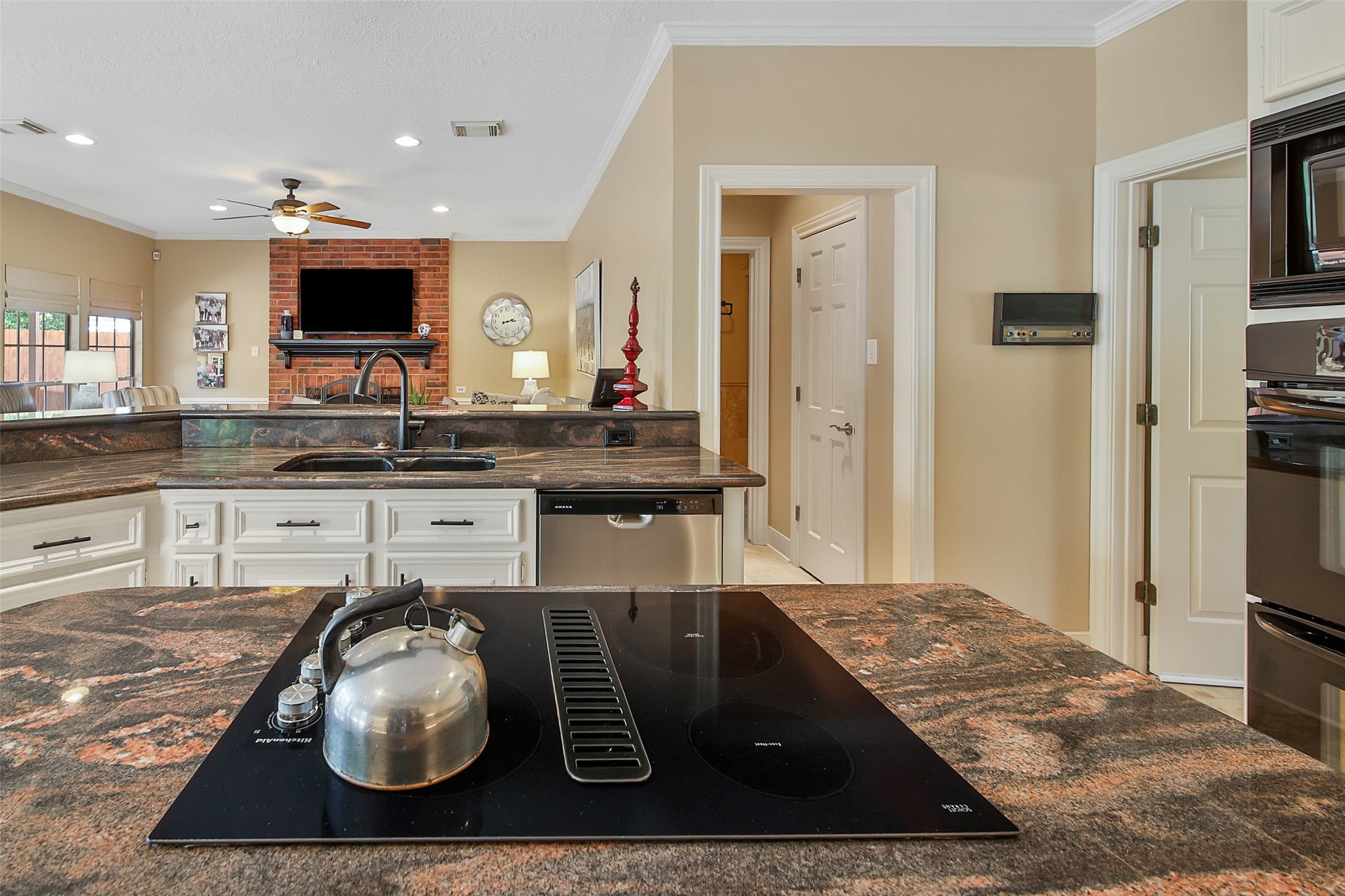 This photo showcases a modern kitchen with granite countertops and a central island featuring a stovetop. The open layout extends to a cozy living area with a brick accent wall, a mounted TV, and a ceiling fan, creating a warm, inviting space.