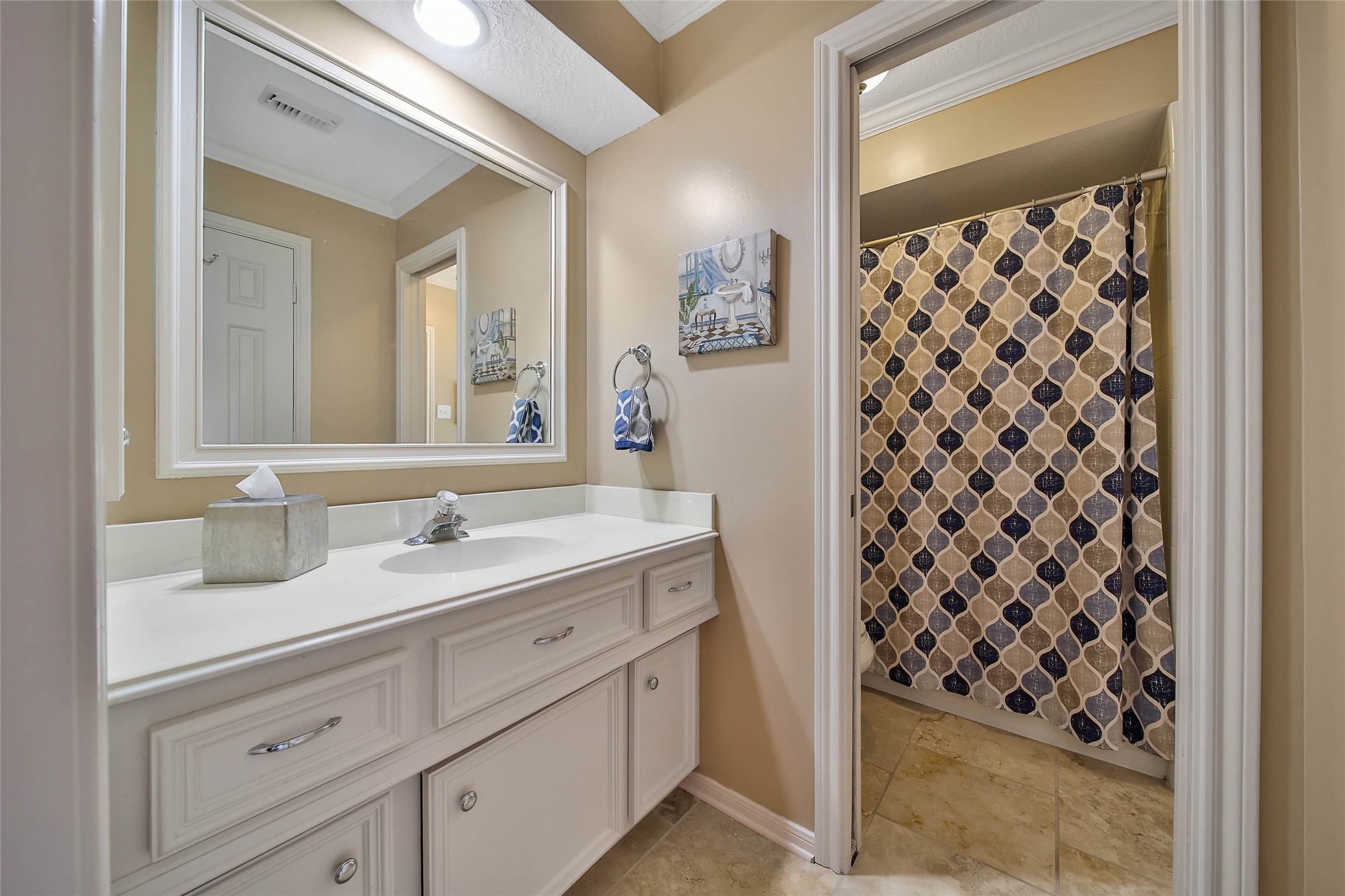 This bathroom features a spacious white vanity with ample storage, a large mirror, and a modern faucet. The shower area is separated by a stylish patterned curtain. Warm beige walls and tile flooring create a cozy atmosphere.