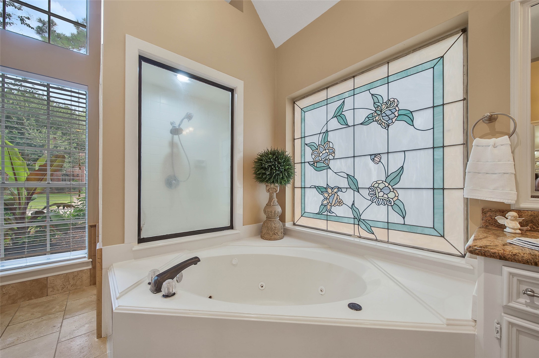 This bathroom features a luxurious corner jacuzzi tub with a stained glass window, providing privacy and style. The space is accented with a frosted glass shower, natural light from large windows, and elegant tile flooring, creating a serene and inviting atmosphere.