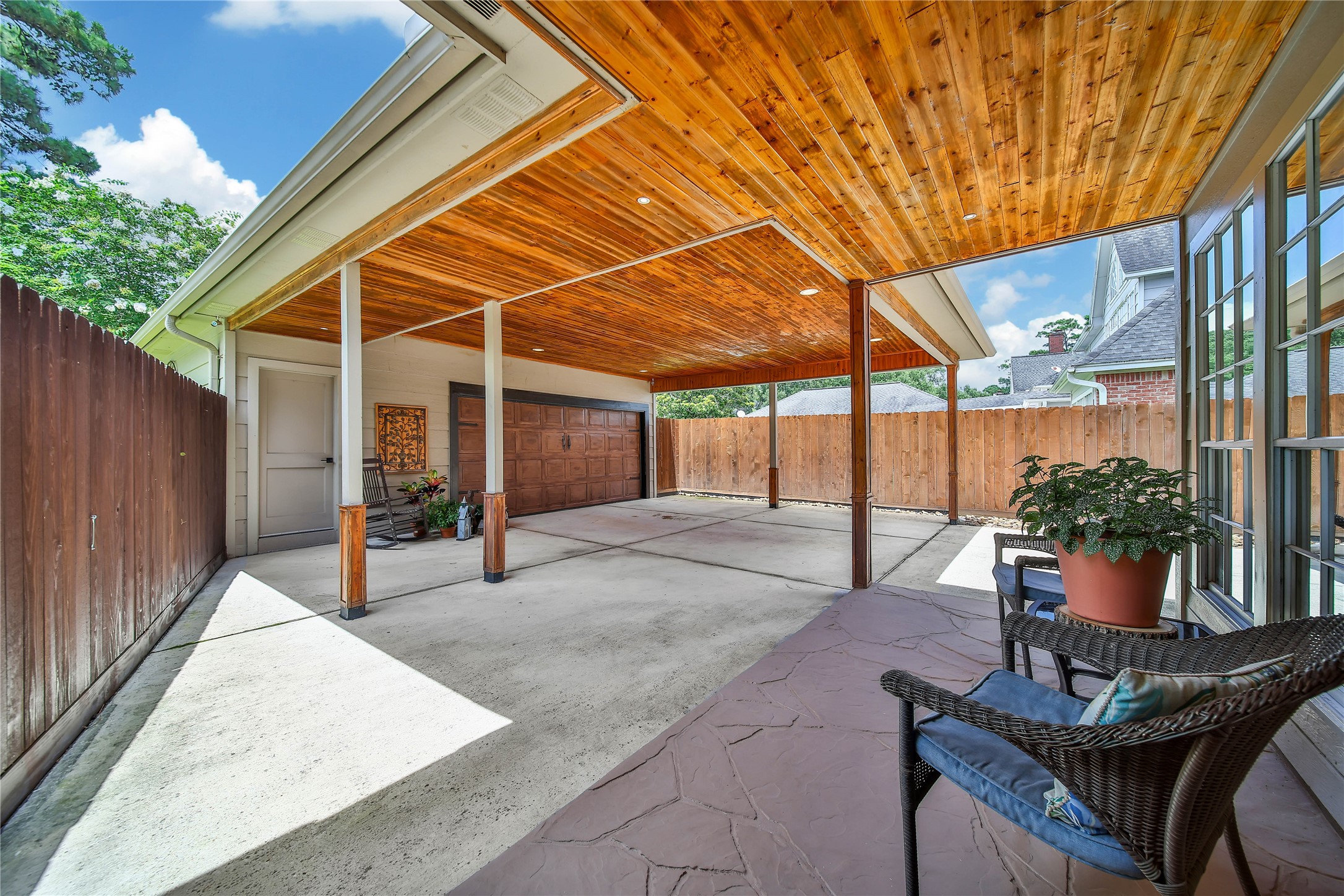 This photo showcases a spacious covered patio with a beautifully finished wood ceiling, adjacent to a garage. The area is partially enclosed by a wooden fence, providing privacy. There's a cozy seating area with a potted plant, perfect for relaxation or entertaining.